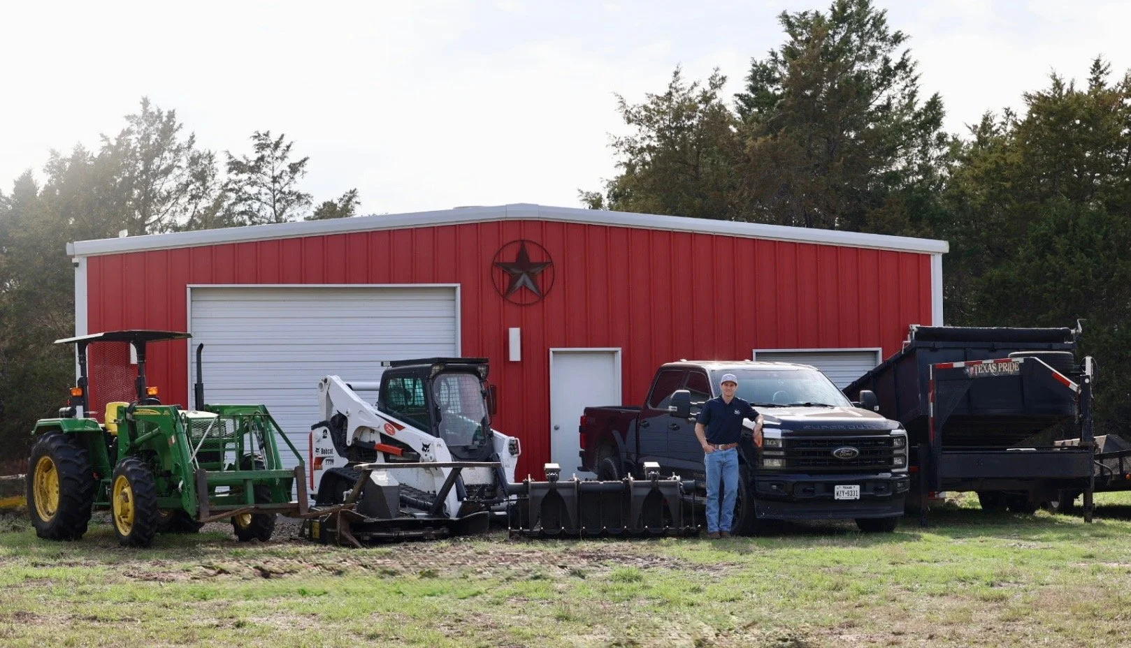 A man standing next to a black pickup truck in front of a red barn with a decorative star on it. The scene includes a green tractor, a white Bobcat skid-steer loader, and a black truck with a trailer. The background has trees and a cloudy sky.