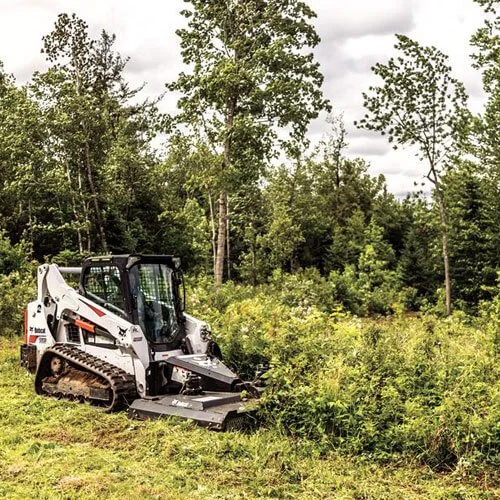 A small white and black skid steer loader working on a lush green overgrown field, surrounded by tall trees under a cloudy sky.