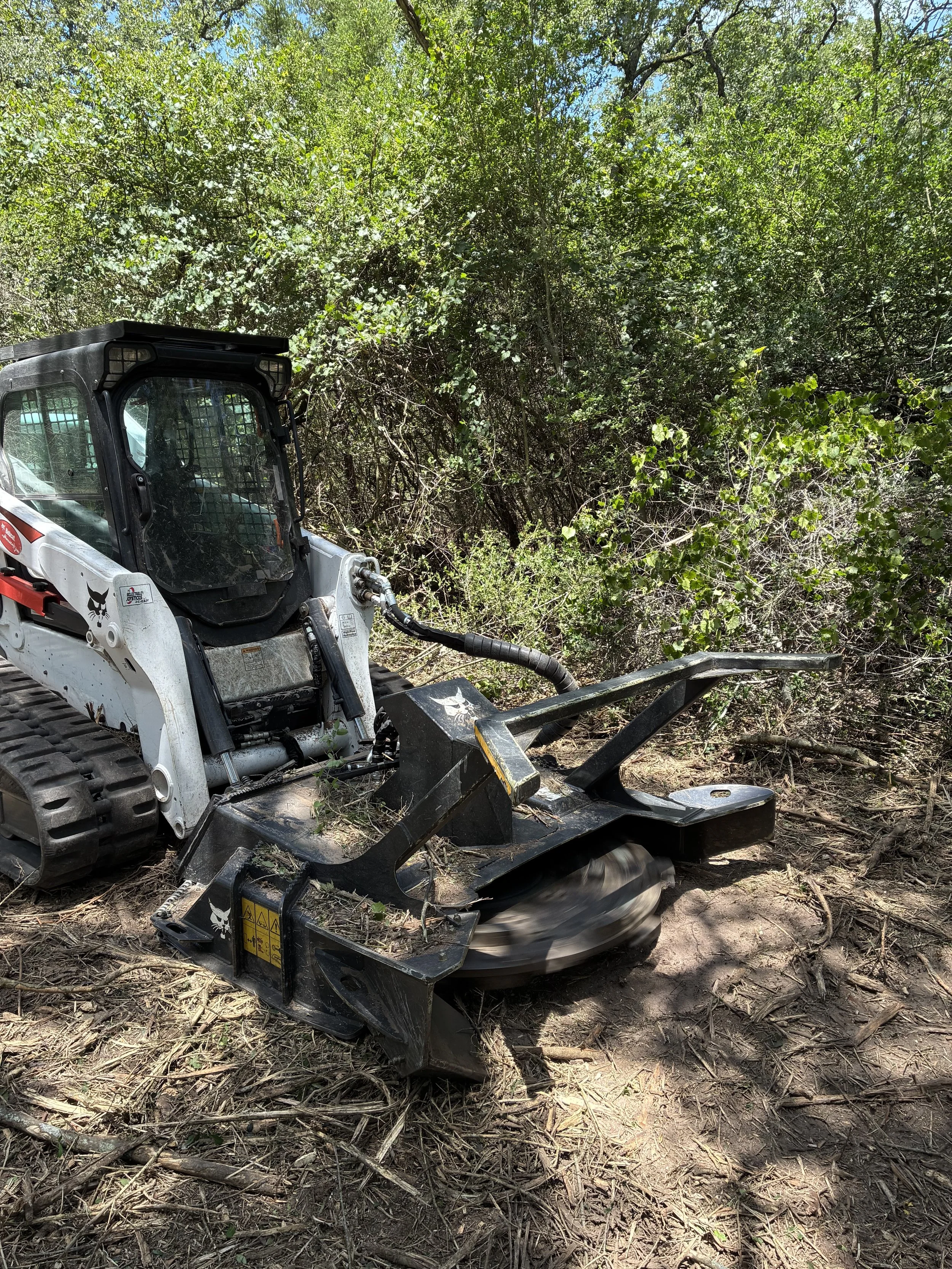 A piece of heavy machinery, specifically a skid steer loader, operating in a wooded area with green foliage and a dirt ground.