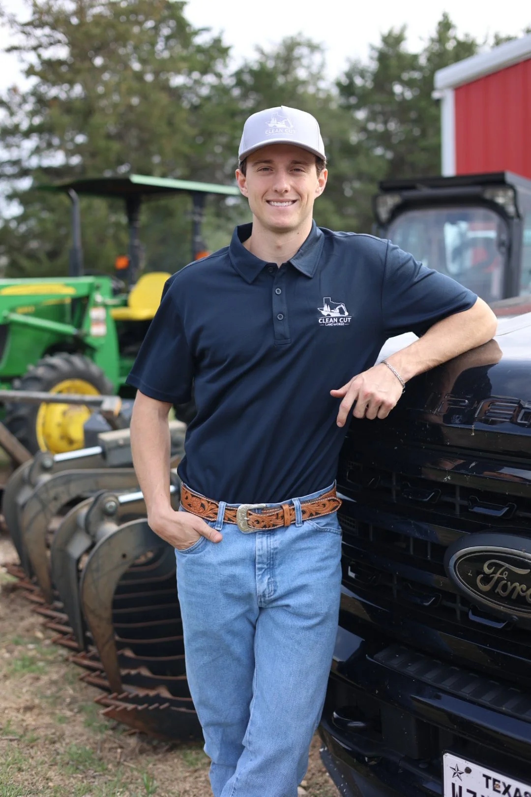 A smiling young man wearing a navy polo shirt and a light gray baseball cap, standing outdoors next to a black Ford vehicle, with farm equipment and trees in the background.