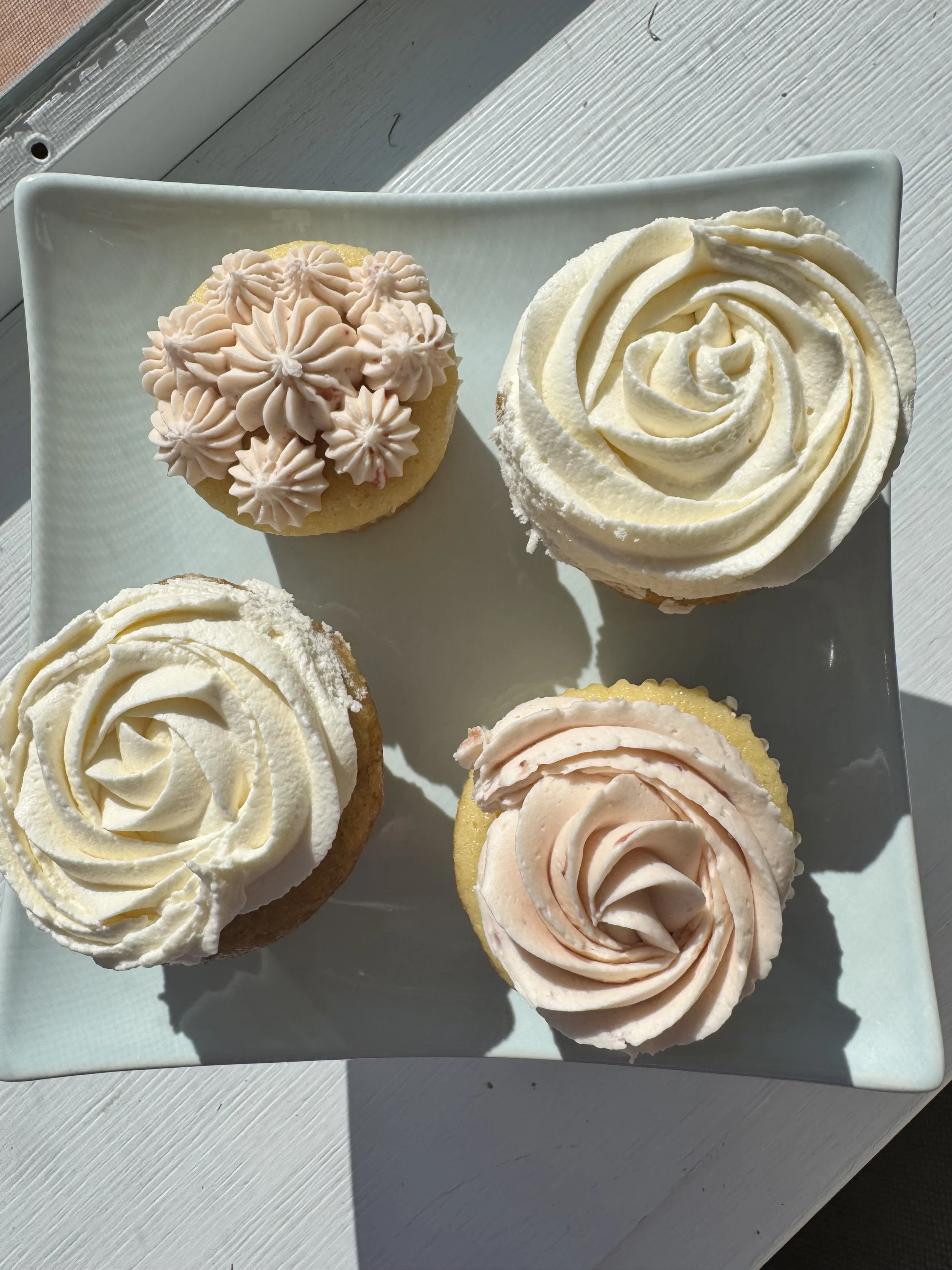 Four cupcakes on a white square plate, decorated with swirls of white and pink icing, some with piped flower patterns.