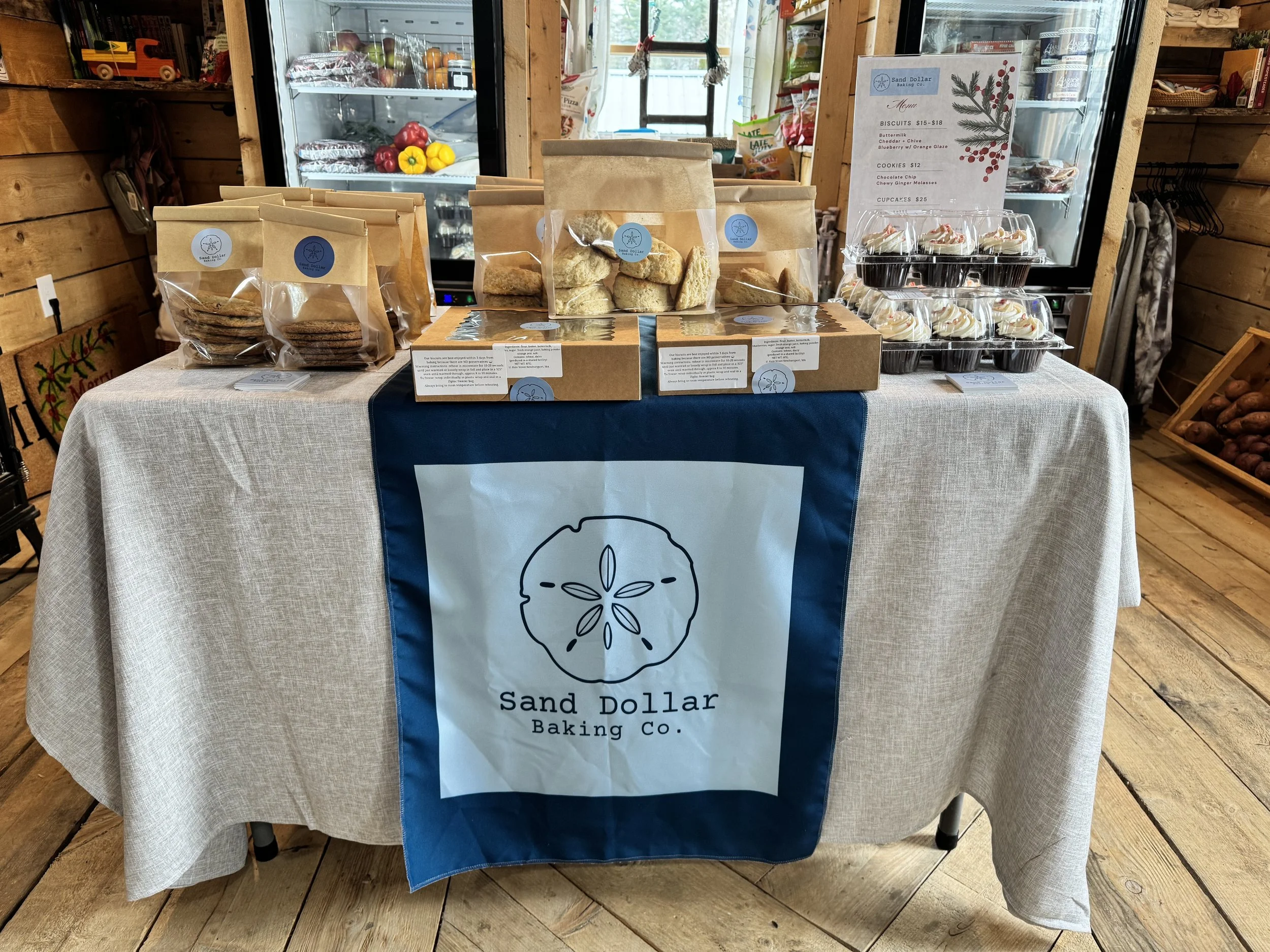 Table display for Sand Dollar Baking Co. with packaged cookies and baked goods, a menu sign, and baked items behind a glass refrigerator, set in a rustic interior with wooden walls and flooring.