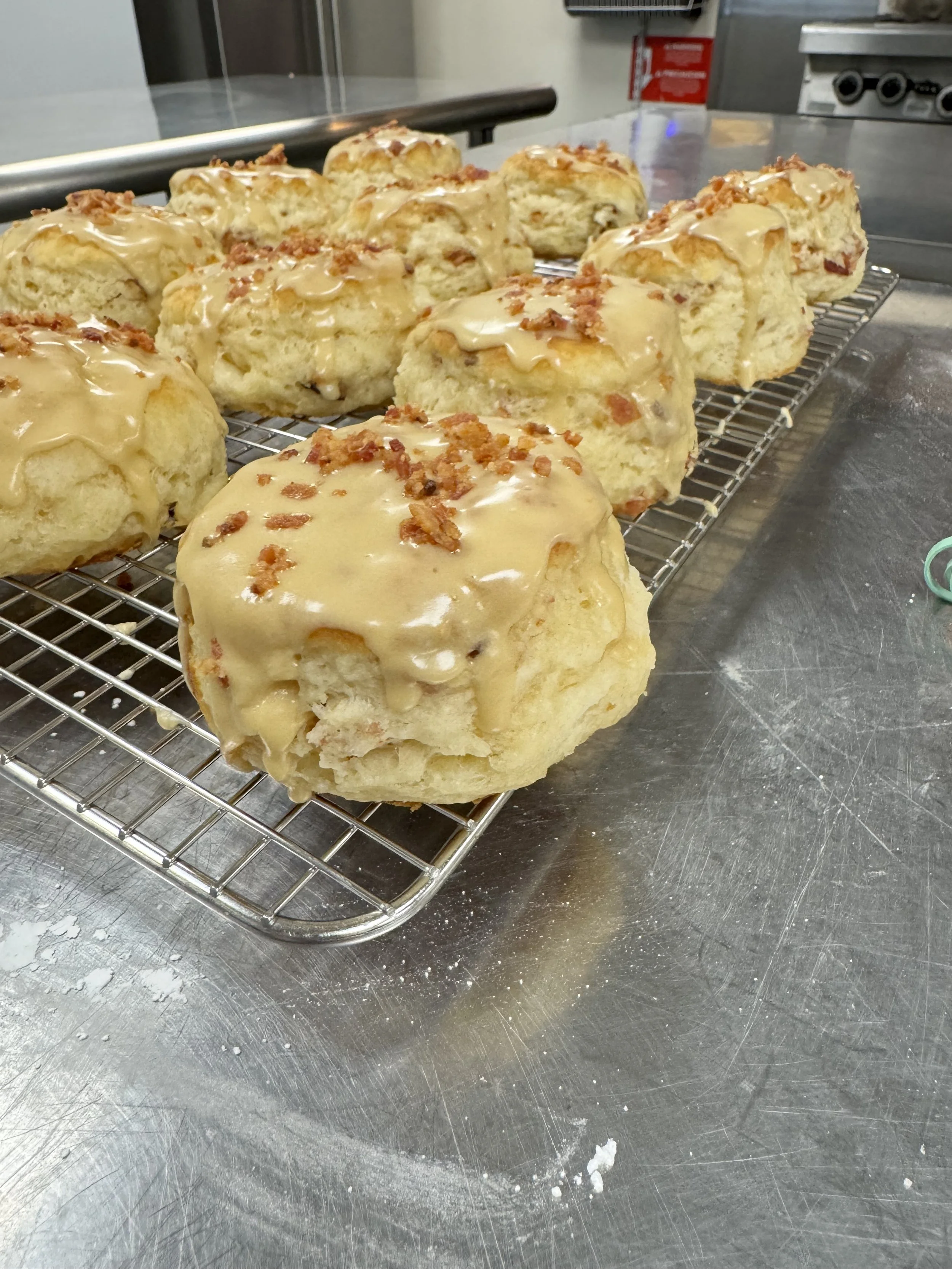 Several glazed scones topped with bacon bits cooling on a wire rack in a commercial kitchen.