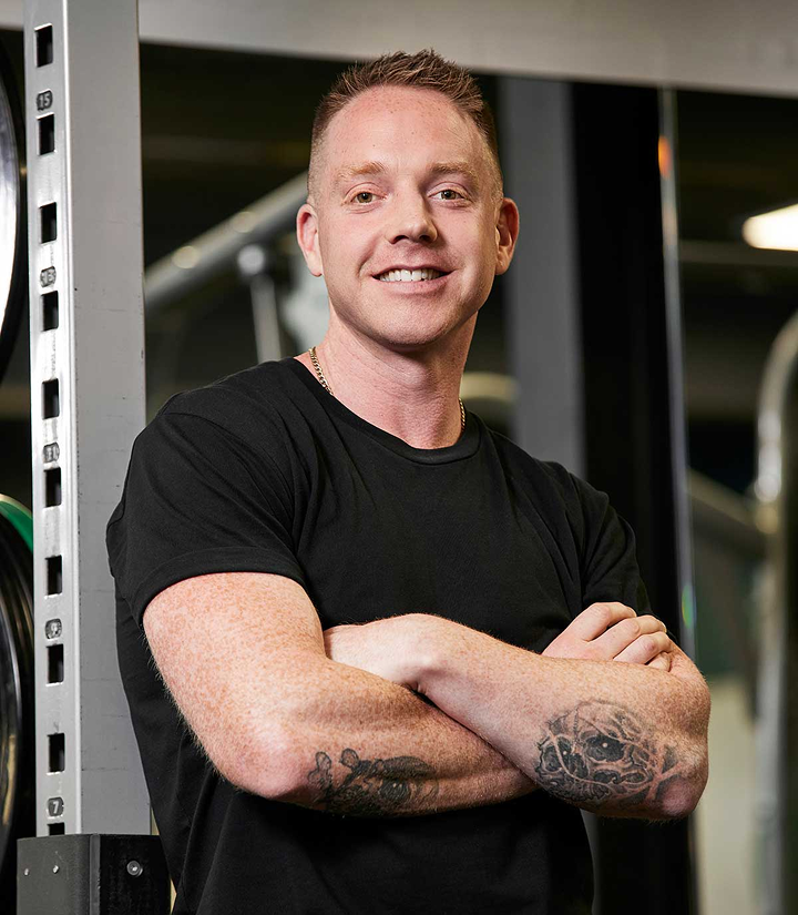 Man with short red hair and tattoos on his arms, wearing a black t-shirt, standing with arms crossed in a gym.
