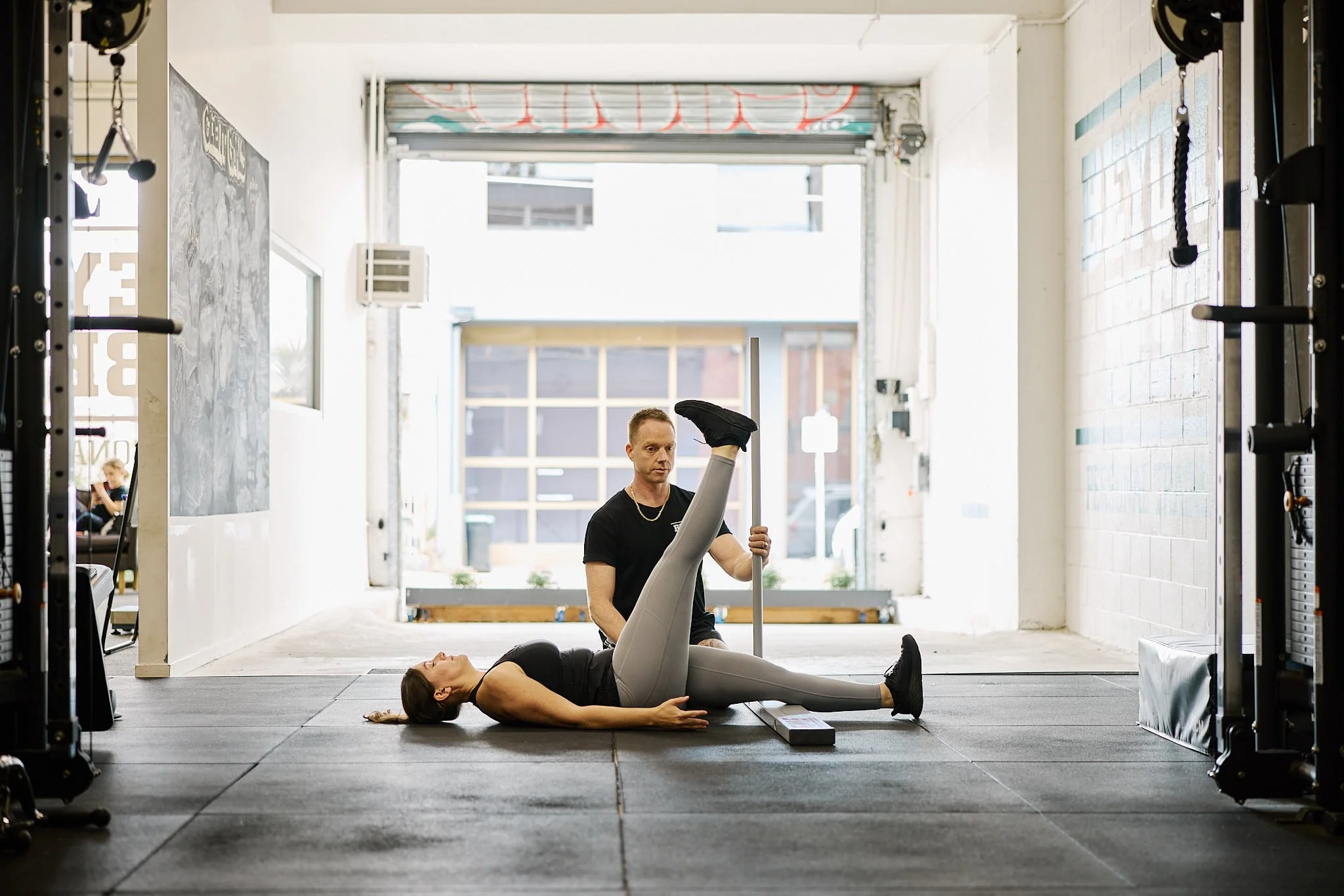 Personal trainer assisting woman with leg stretch on gym floor during workout session.