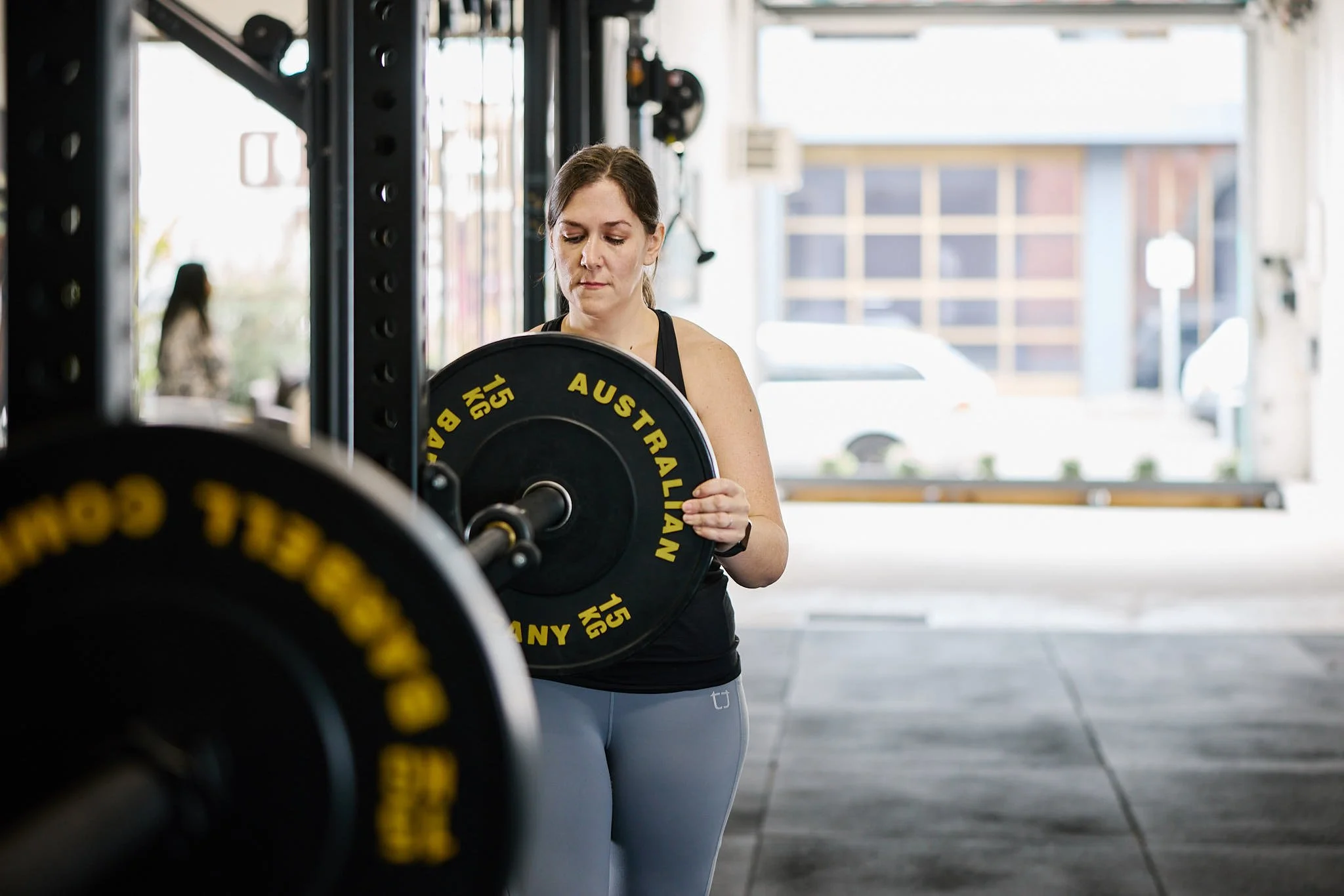 A man with a beard, tattoos, and a bald head bending over to lift a red Our Good PROUD barbell with 25 kg weights in a gym. The gym has black flooring and bright overhead lights, with other people working out in the background.