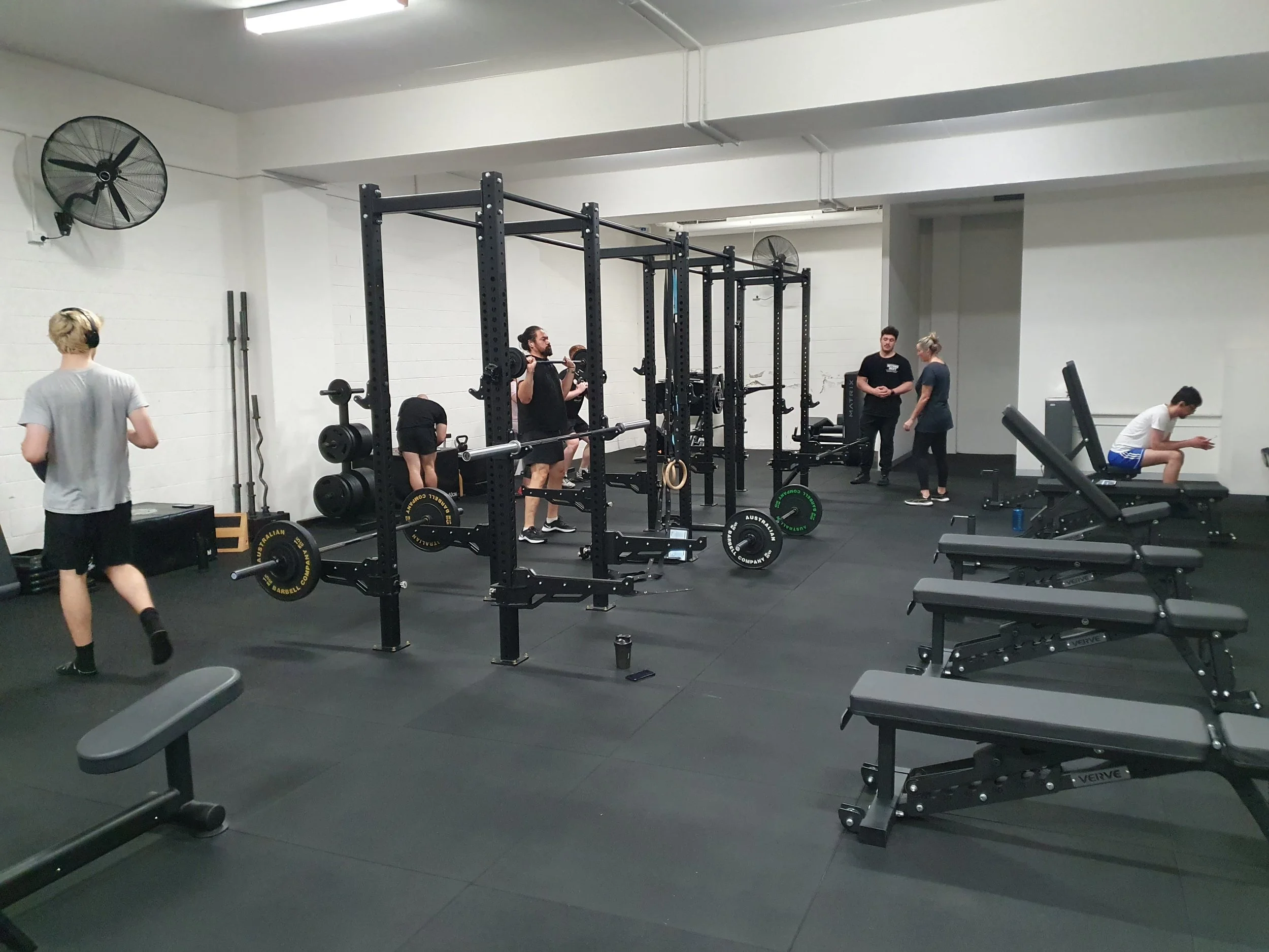 Interior of a spacious gym with workout equipment including benches, weights, and cardio machines, under a high ceiling with skylights.