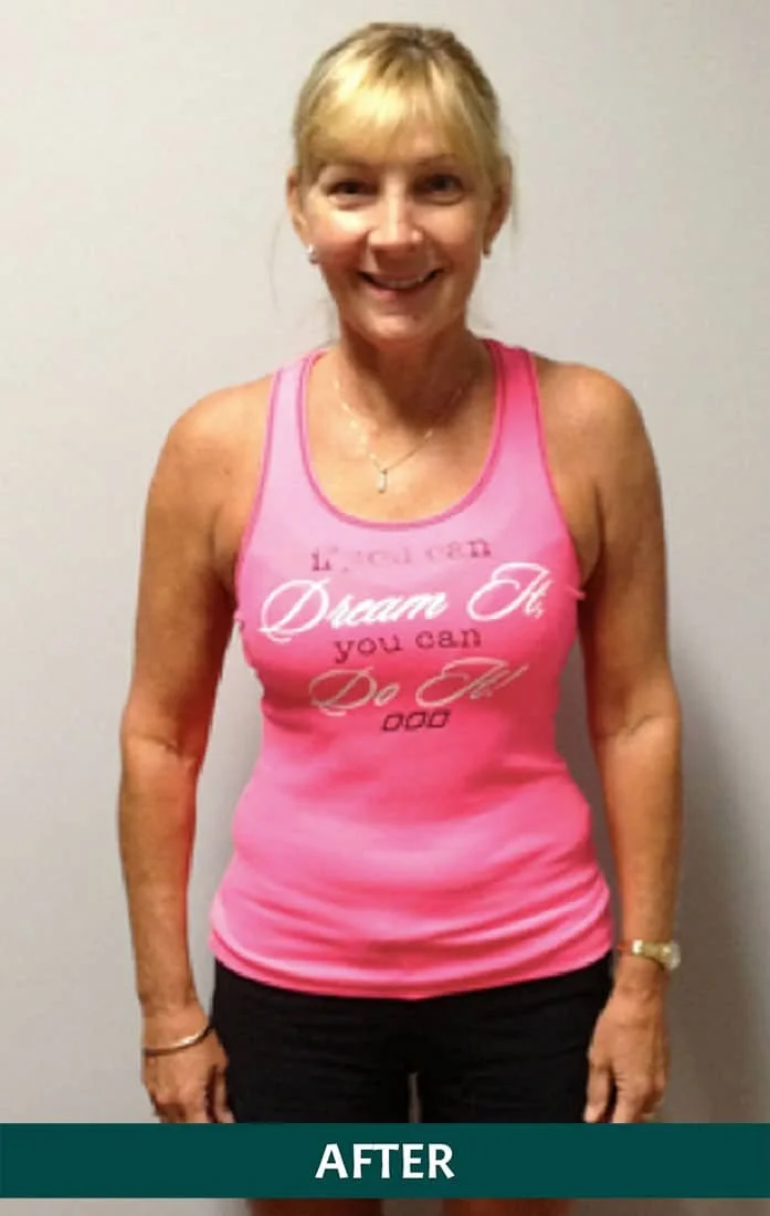 A smiling middle-aged woman with blonde hair, wearing a pink workout tank top with motivational text, standing against a plain wall.
