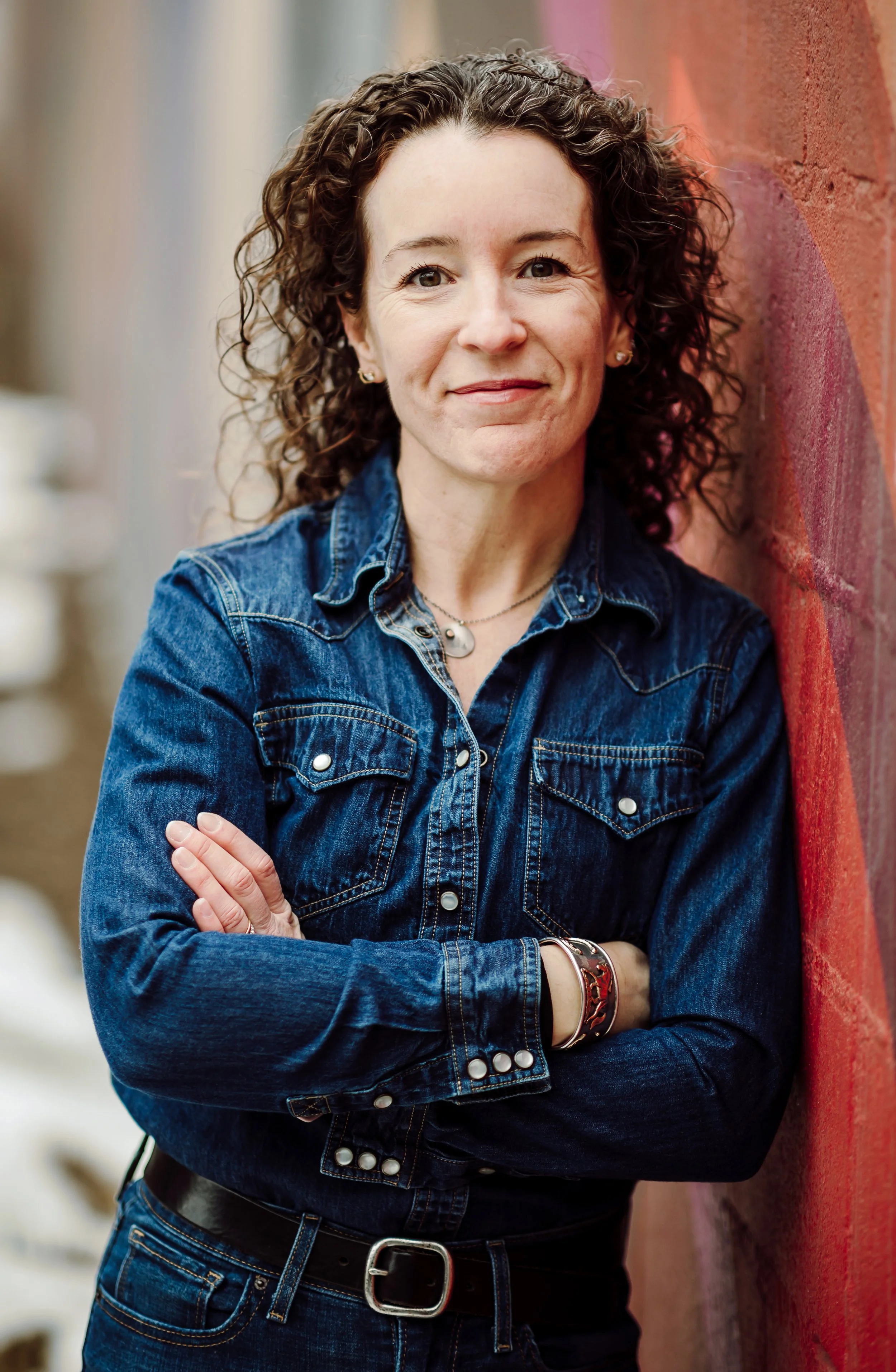 A woman with curly brown hair wearing a denim jacket, standing with arms crossed against a red brick wall.