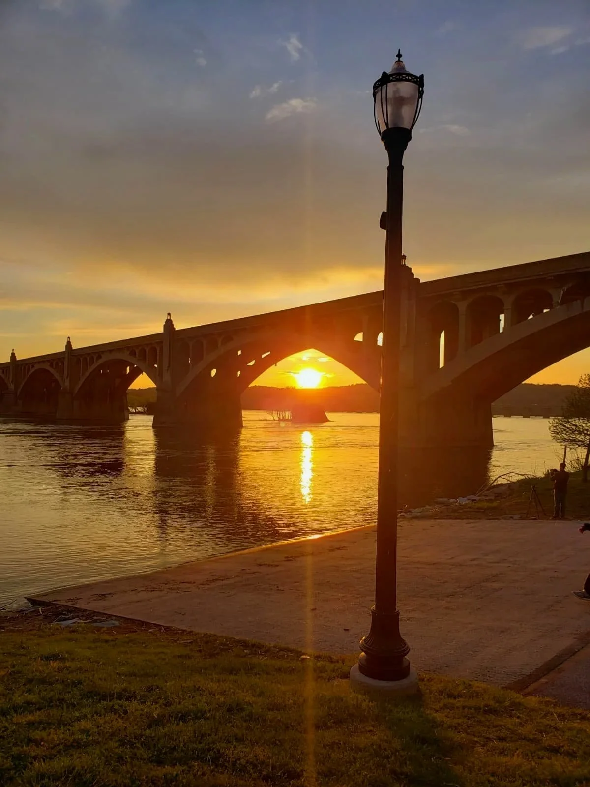 A bridge over a body of water during sunset, with a lamp post in the foreground and a person fishing on the right side.