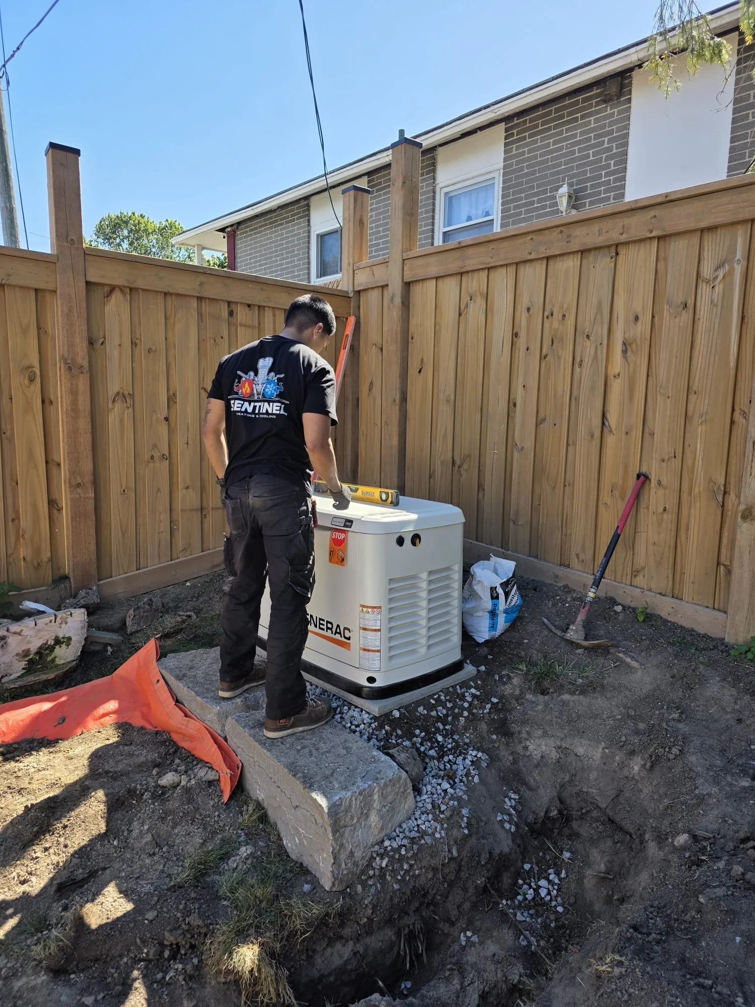 A man working outdoors next to a wooden fence, installing or maintaining a generators with tools nearby.
