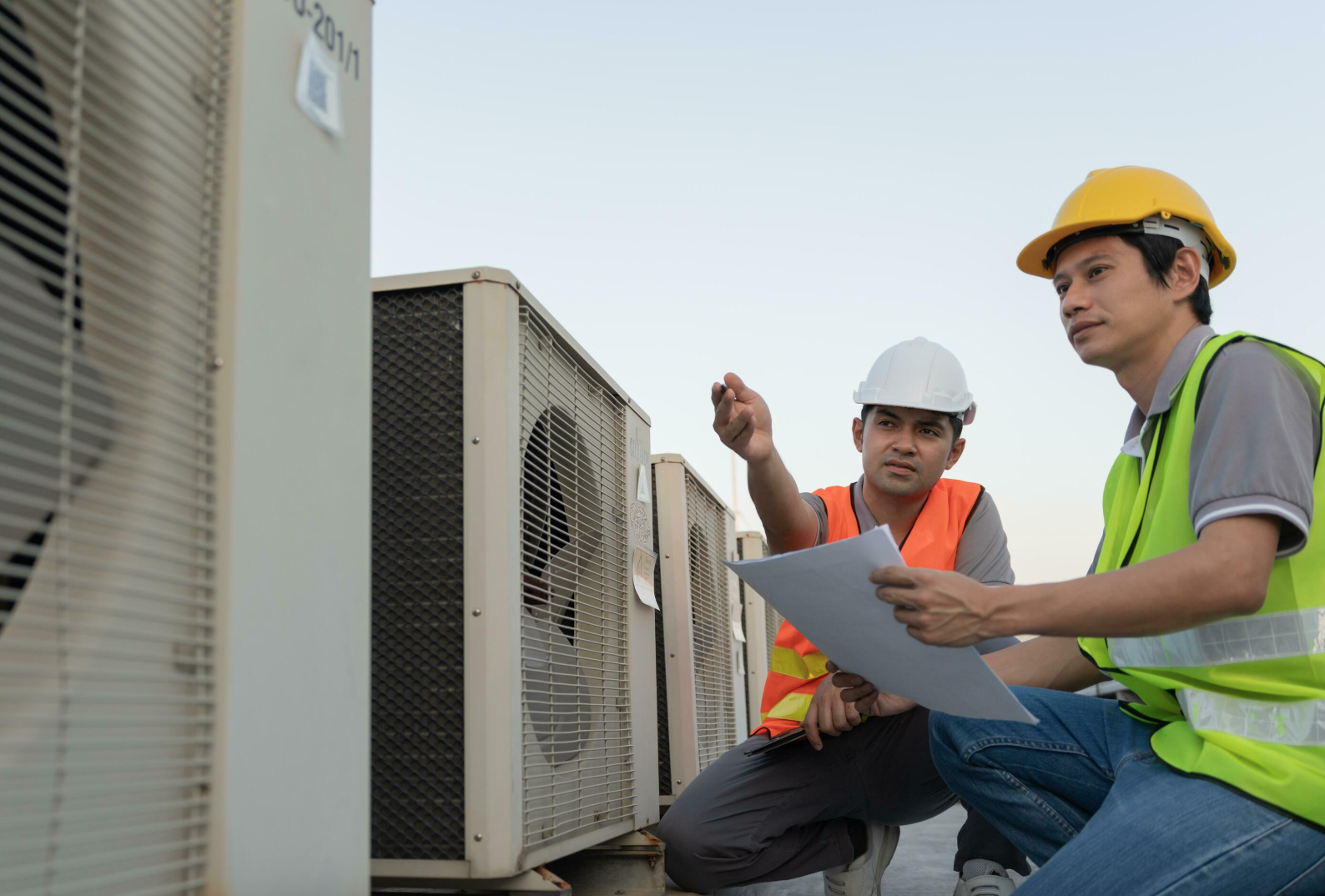 Two male technicians wearing safety gear inspecting large industrial air conditioning units outside.
