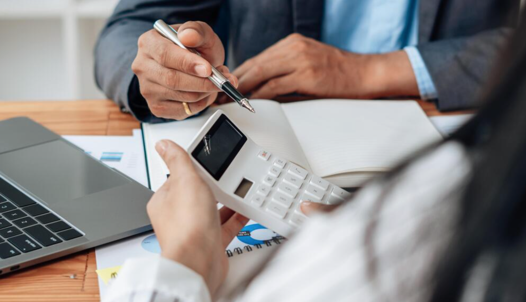 Two people working at a desk with a laptop, calculator, and documents; one is holding a calculator, and the other is pointing with a pen.