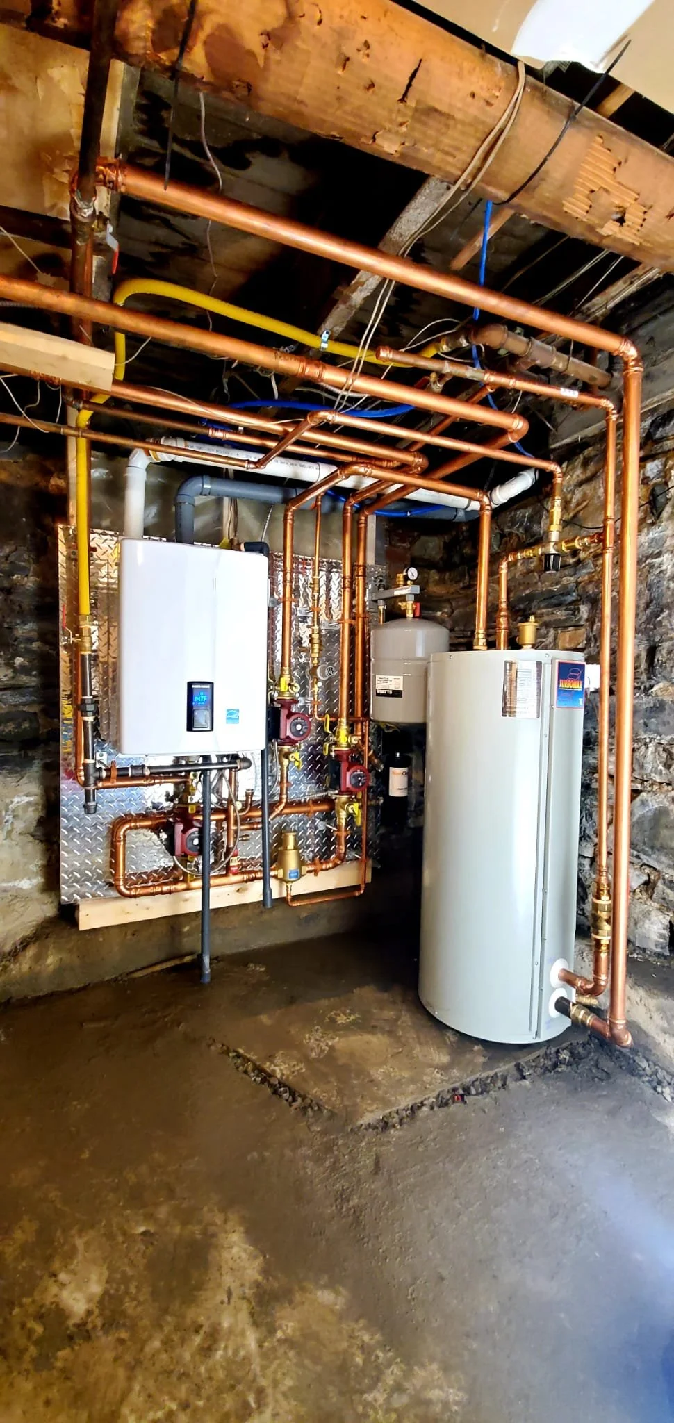 A utility room with copper pipes, a hot water heater tank, and various plumbing and heating components mounted on a diamond plate wall, with a rough concrete floor.