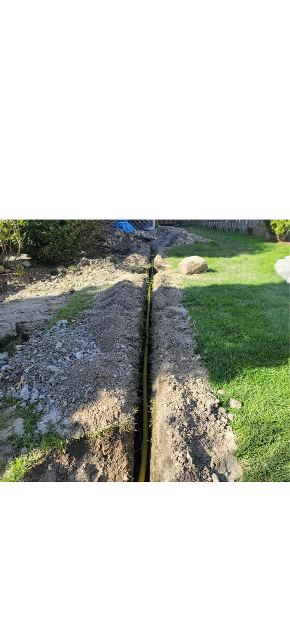 A freshly dug trench in a backyard with a yellow conduit pipe running through it, surrounded by grass, soil, and some landscaping rocks.