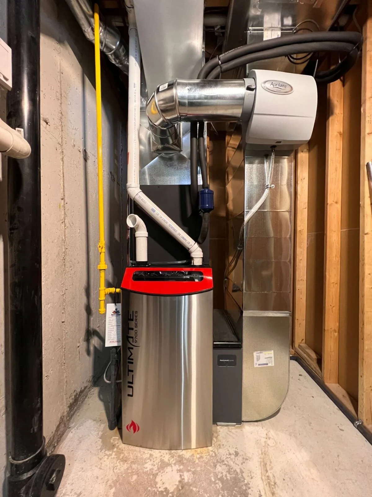 This image shows a basement utility area with a gray furnace, a stainless steel water softener labeled 'ULTIMATE', pipes, a yellow gas line, and ductwork.