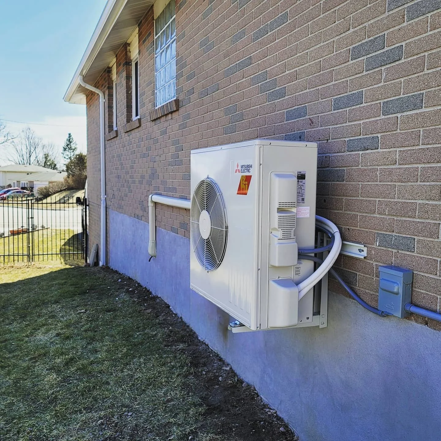 Outdoor view of an air conditioning unit mounted on a brick wall of a house, with a grassy yard and neighboring houses in the background.