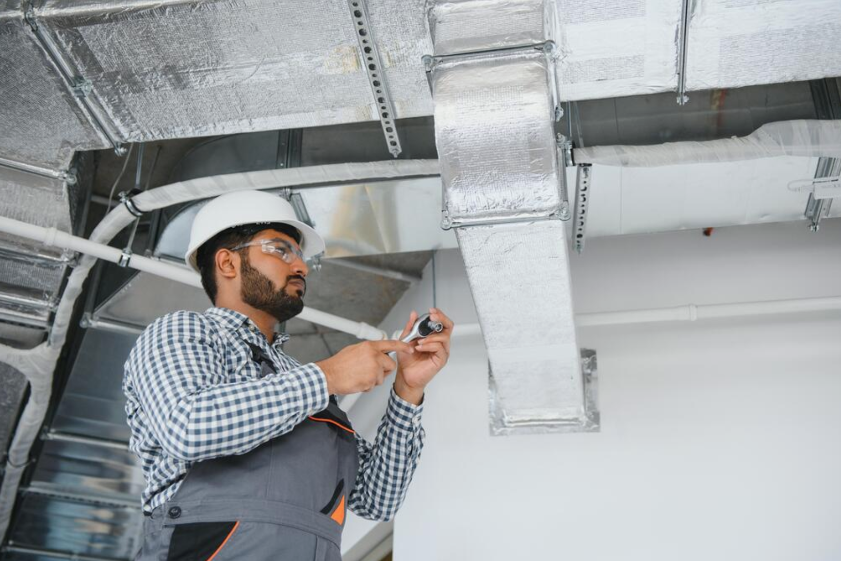 A man wearing a white safety helmet, safety glasses, and a checkered shirt inspects HVAC ductwork with a handheld device in a commercial or industrial setting.