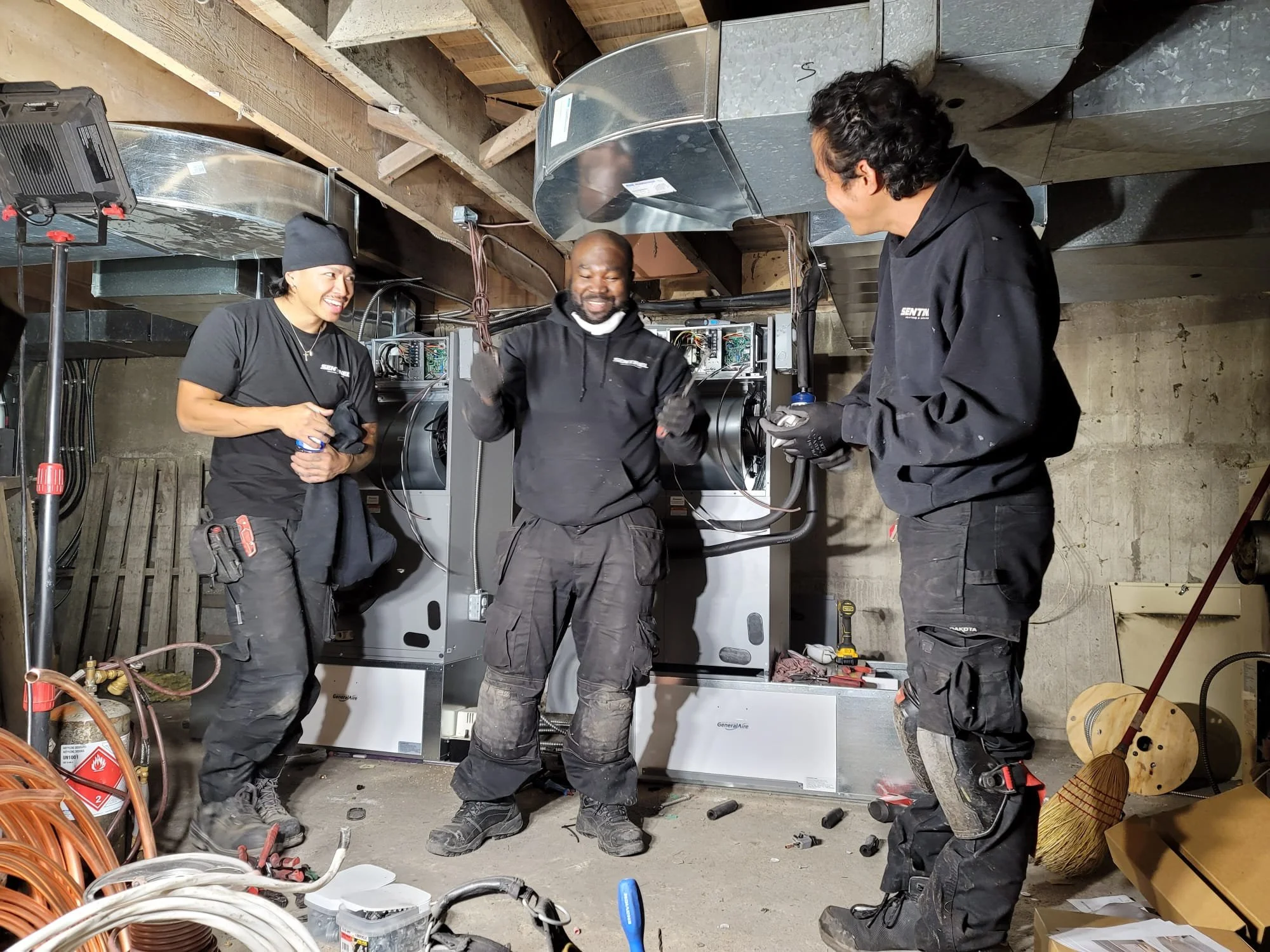 Three construction workers are smiling and chatting in a basement. They are wearing black work clothes, and tools are scattered around them. One worker is gesturing with his fists, while the others listen.