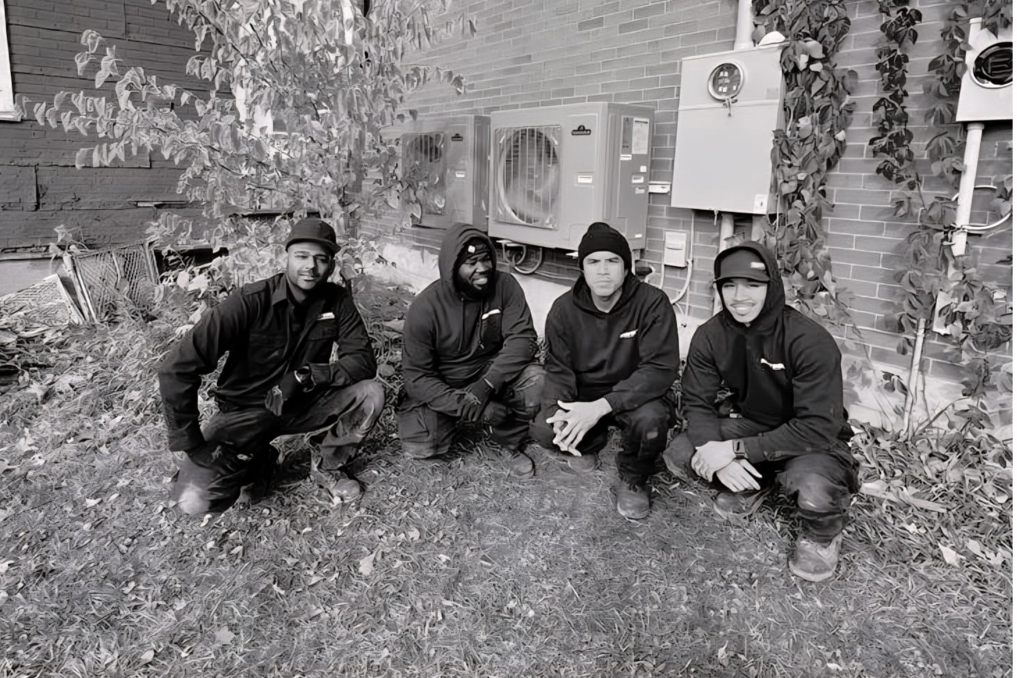 Four workers wearing dark uniforms kneeling on the grass in front of a brick wall with utility boxes and plants.