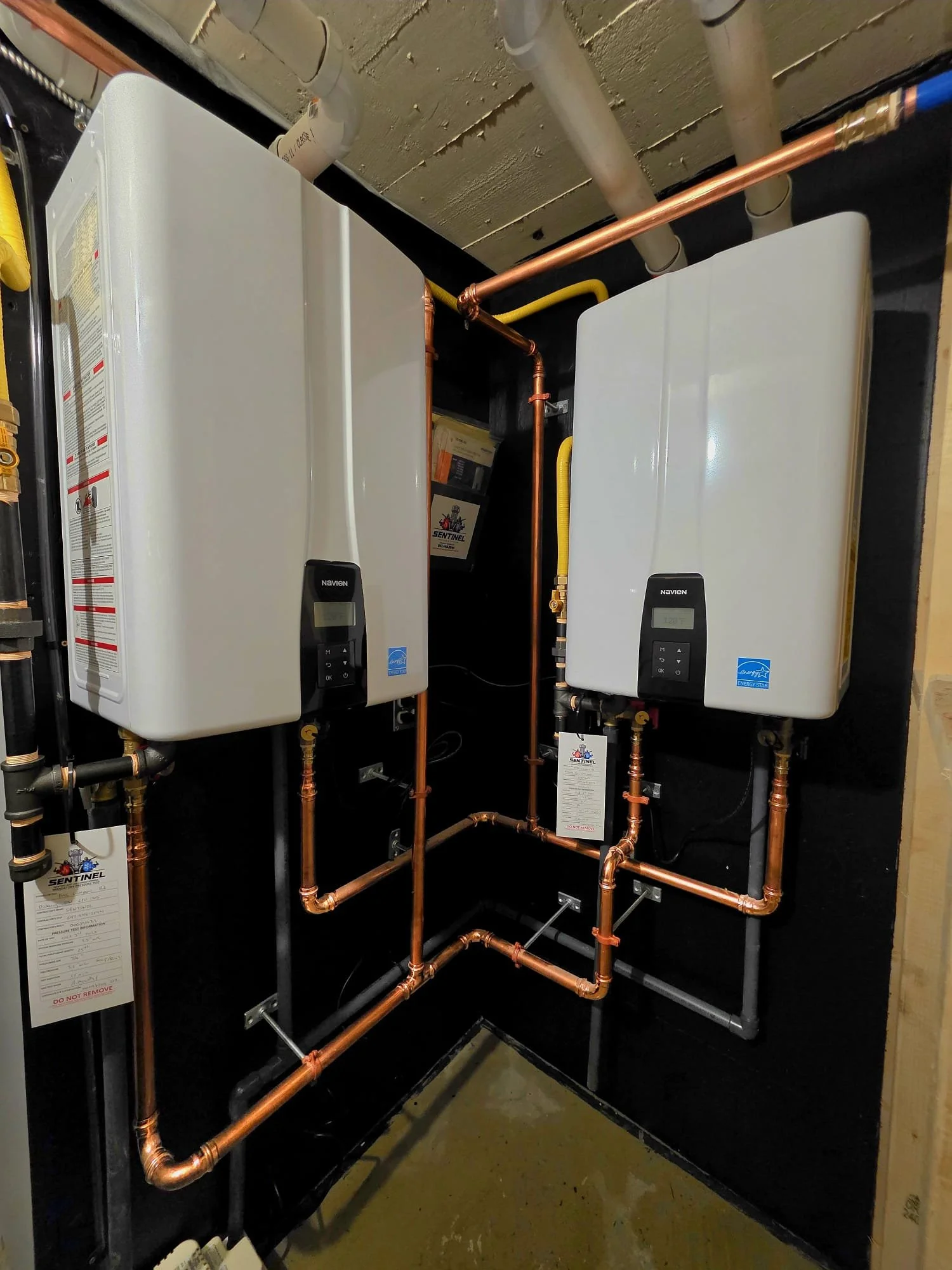 Two white tankless water heaters with digital displays mounted on a black wall, connected by copper and black pipes in a utility room.