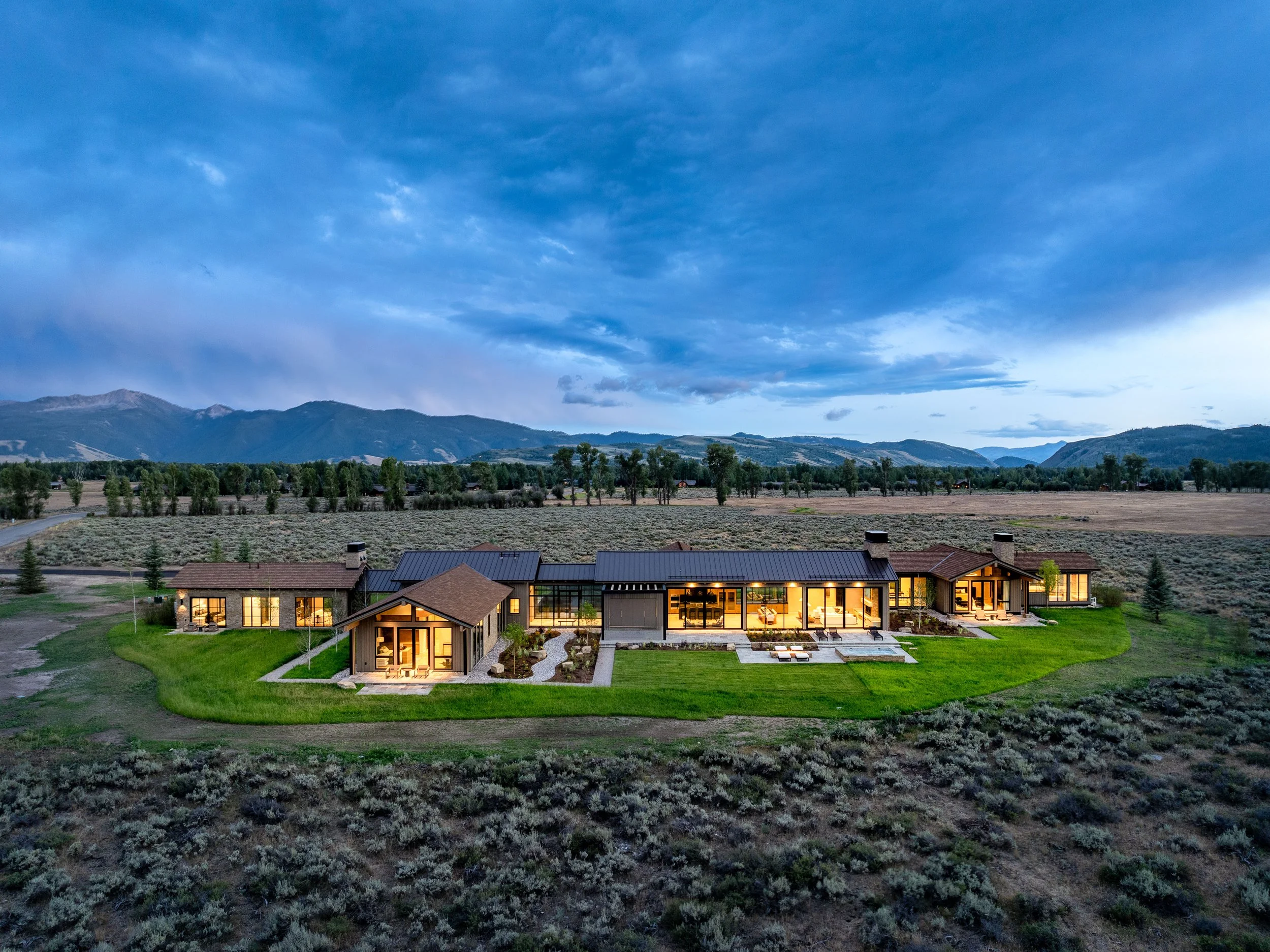 An aerial view of a modern house at dusk with illuminated interior, surrounded by open fields and mountains in the background.