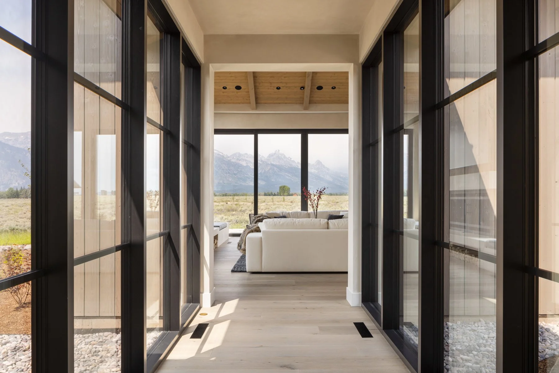 View through glass doors into a bright living room with white sofas, large windows showing mountains and open fields, and wooden ceiling beams.