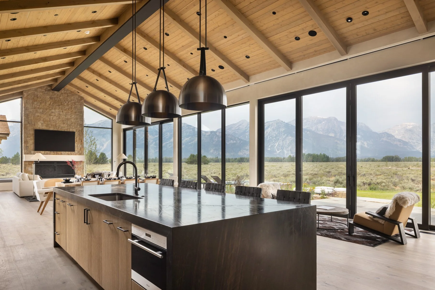 Modern open-concept kitchen and living area with large windows overlooking mountain landscape, wooden ceiling with exposed beams, black pendant lights over kitchen island, light hardwood flooring, and contemporary furniture.