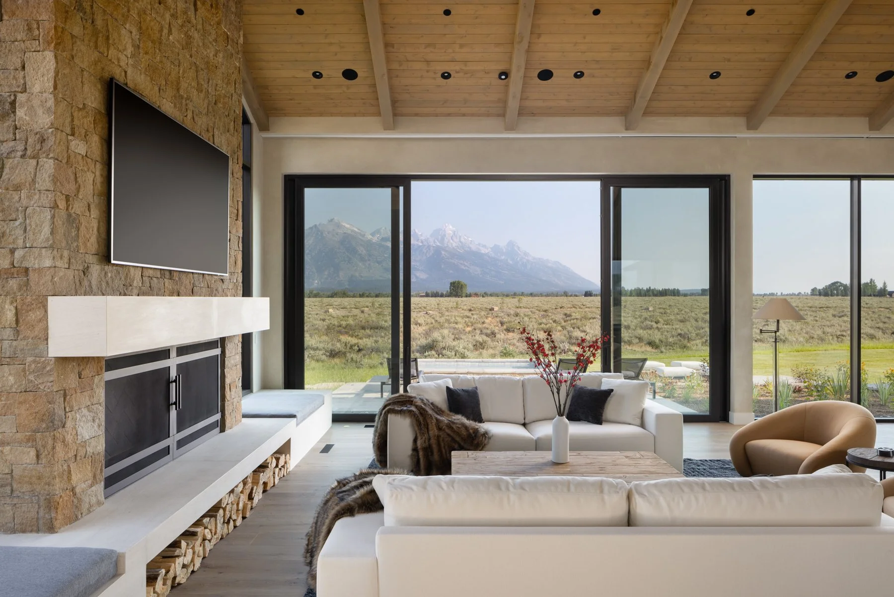 Modern living room with a stone fireplace, a wall-mounted TV, and large sliding glass doors showing a mountain landscape outside. The room has white sofas, a beige chair, a wooden coffee table with a vase of red flowers, and a view of a patio and open field.