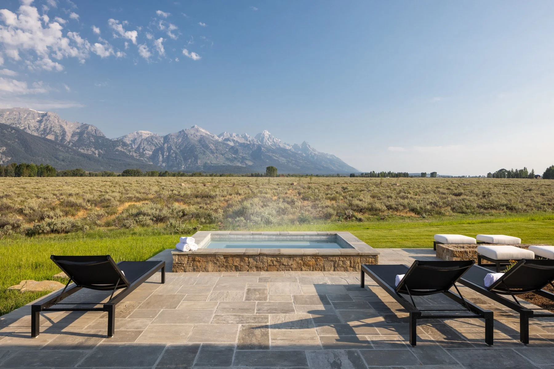 Outdoor hot tub surrounded by lounge chairs and towels, with grassy fields and mountains in the background under a cloudy blue sky.