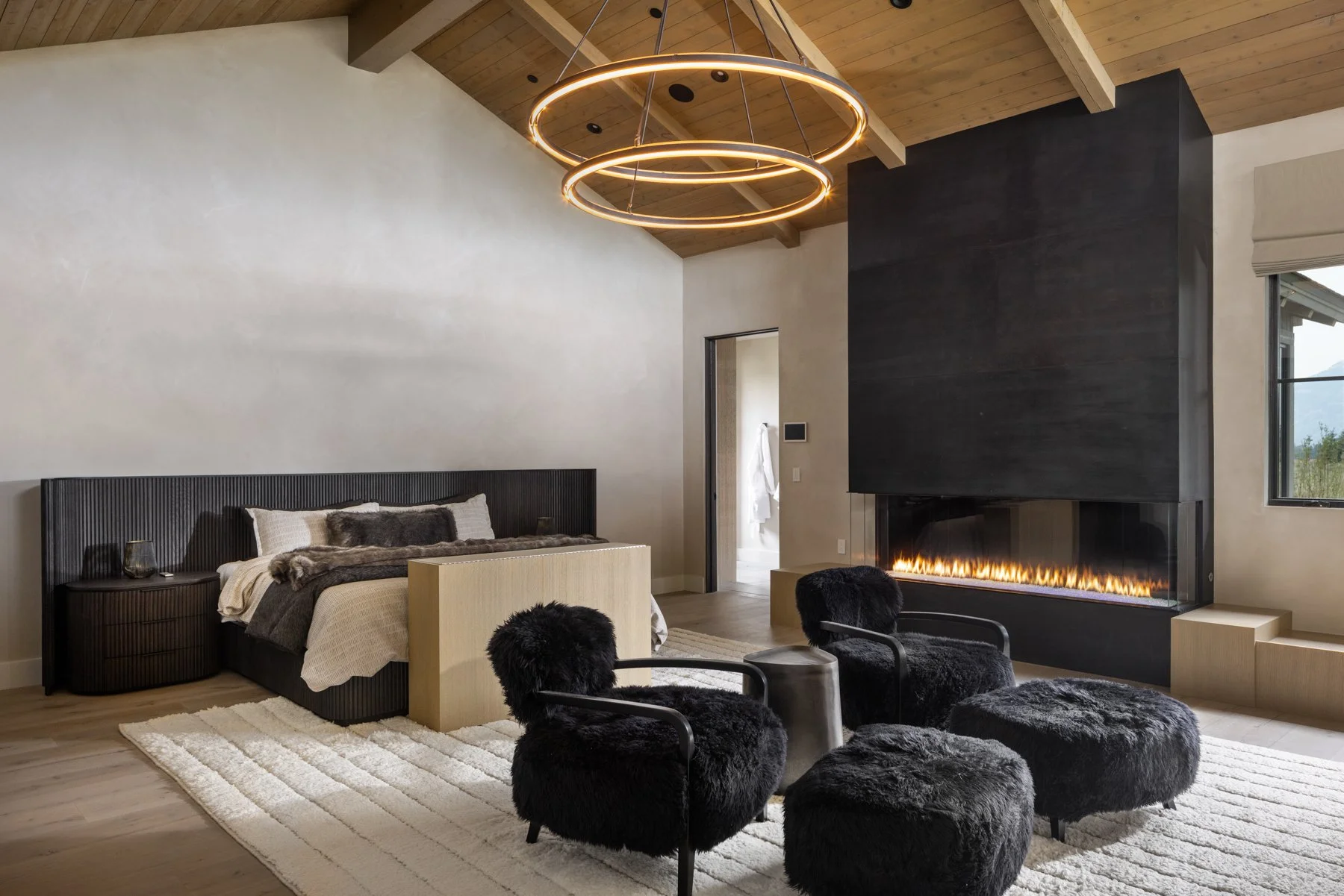 Modern bedroom with a black bed, two black furry armchairs and ottomans, a fireplace with a black wall and glass front, a white textured rug, and a wooden ceiling with circular light fixtures.