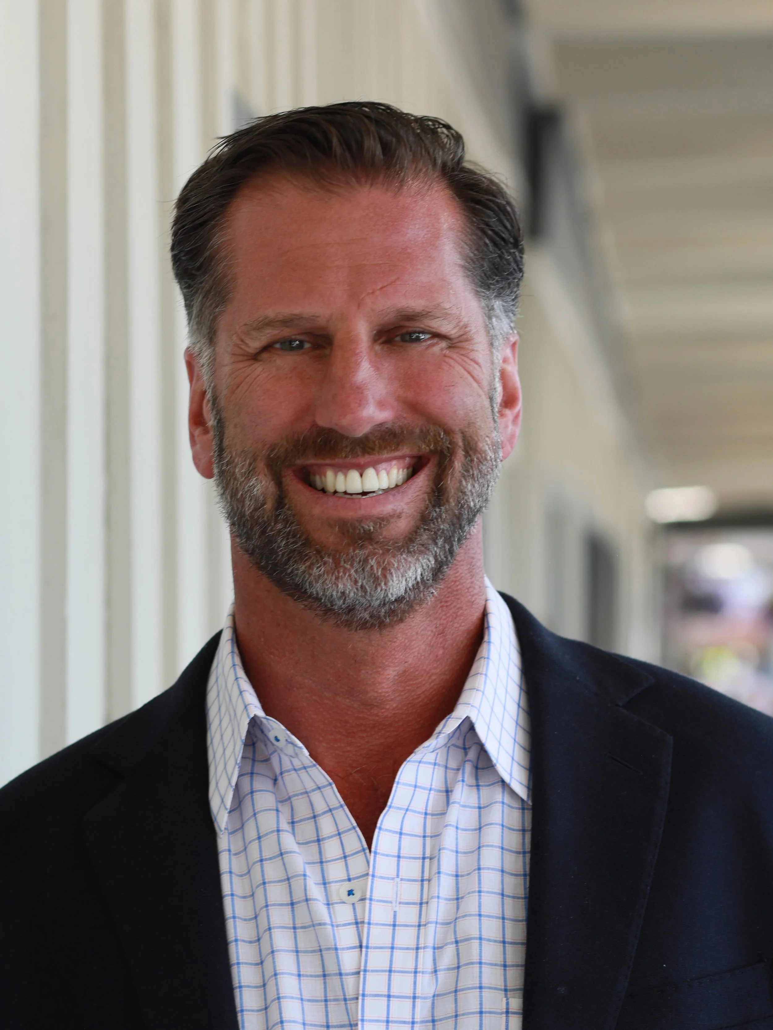 A smiling man with gray hair and a beard wearing a dark blazer and a light blue checked shirt.