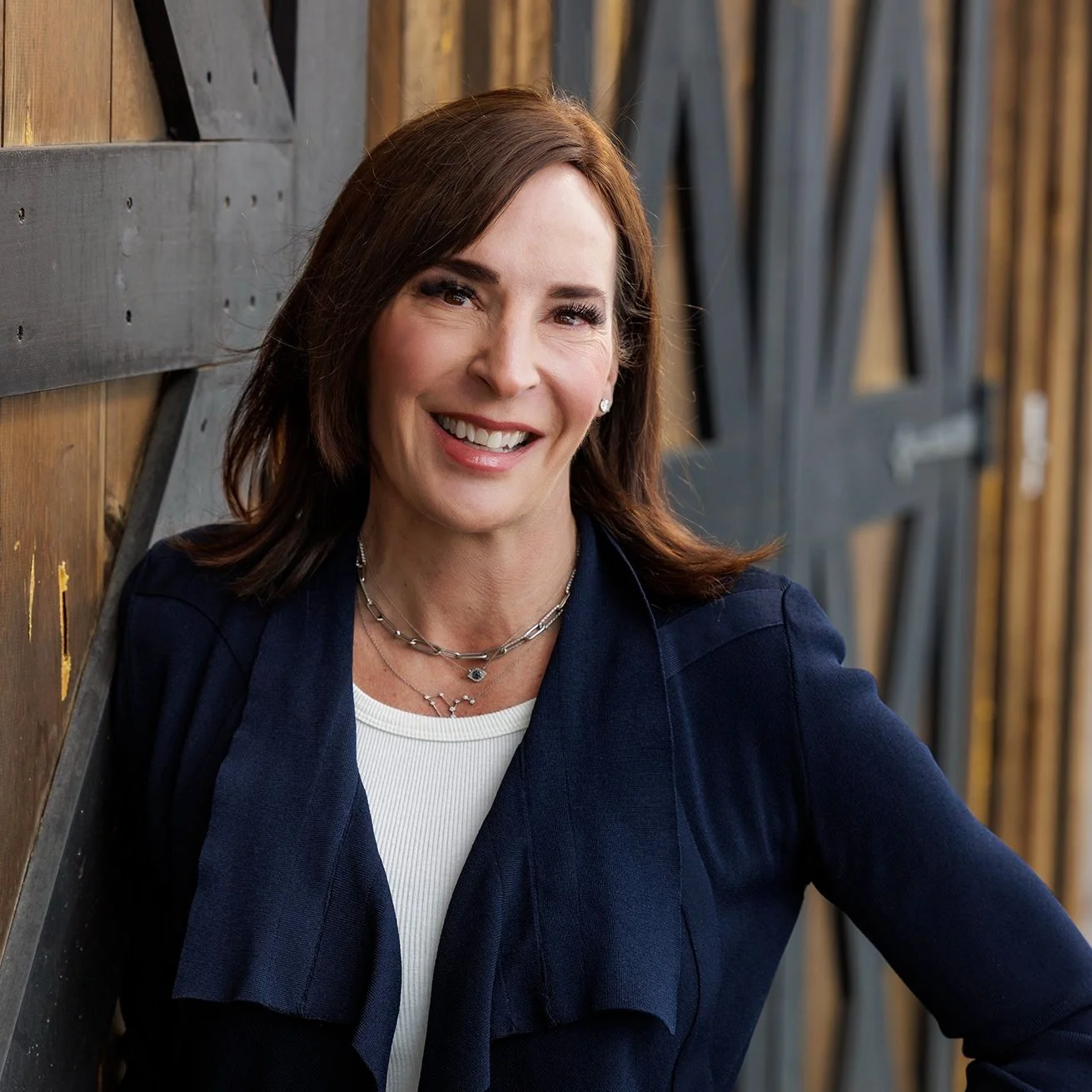 A woman with brown hair smiling, wearing a navy blue jacket and jewelry, standing against a wooden wall with black metal accents.
