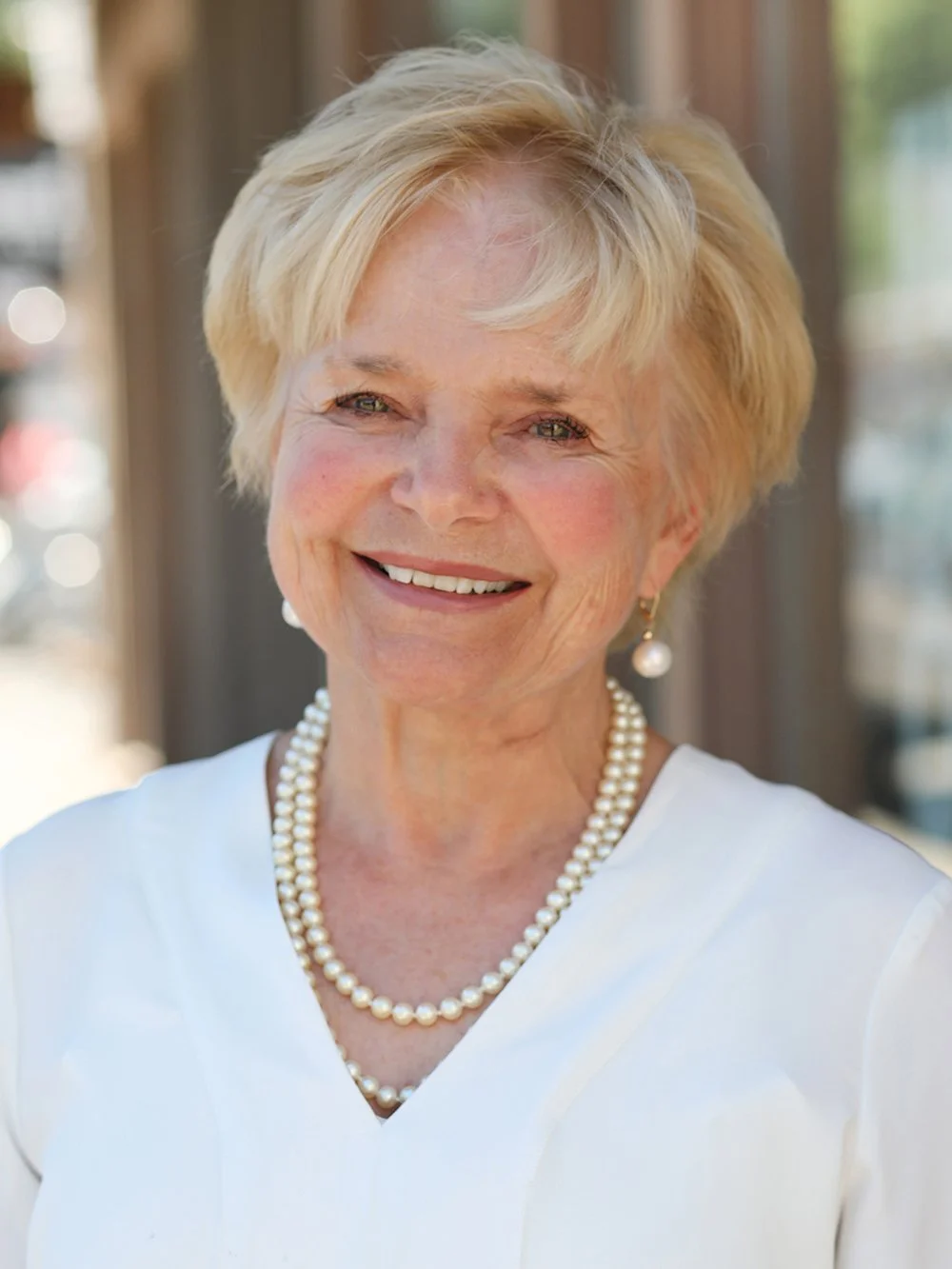 A smiling elderly woman with short blonde hair, wearing pearl earrings and a pearl necklace, dressed in a white top, standing outdoors with a blurred background.