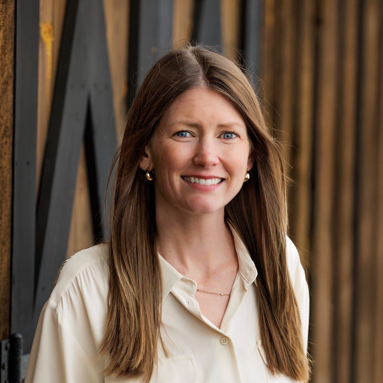 A smiling woman with long brown hair, wearing a cream-colored blouse and gold earrings, standing in front of a rustic wooden background.