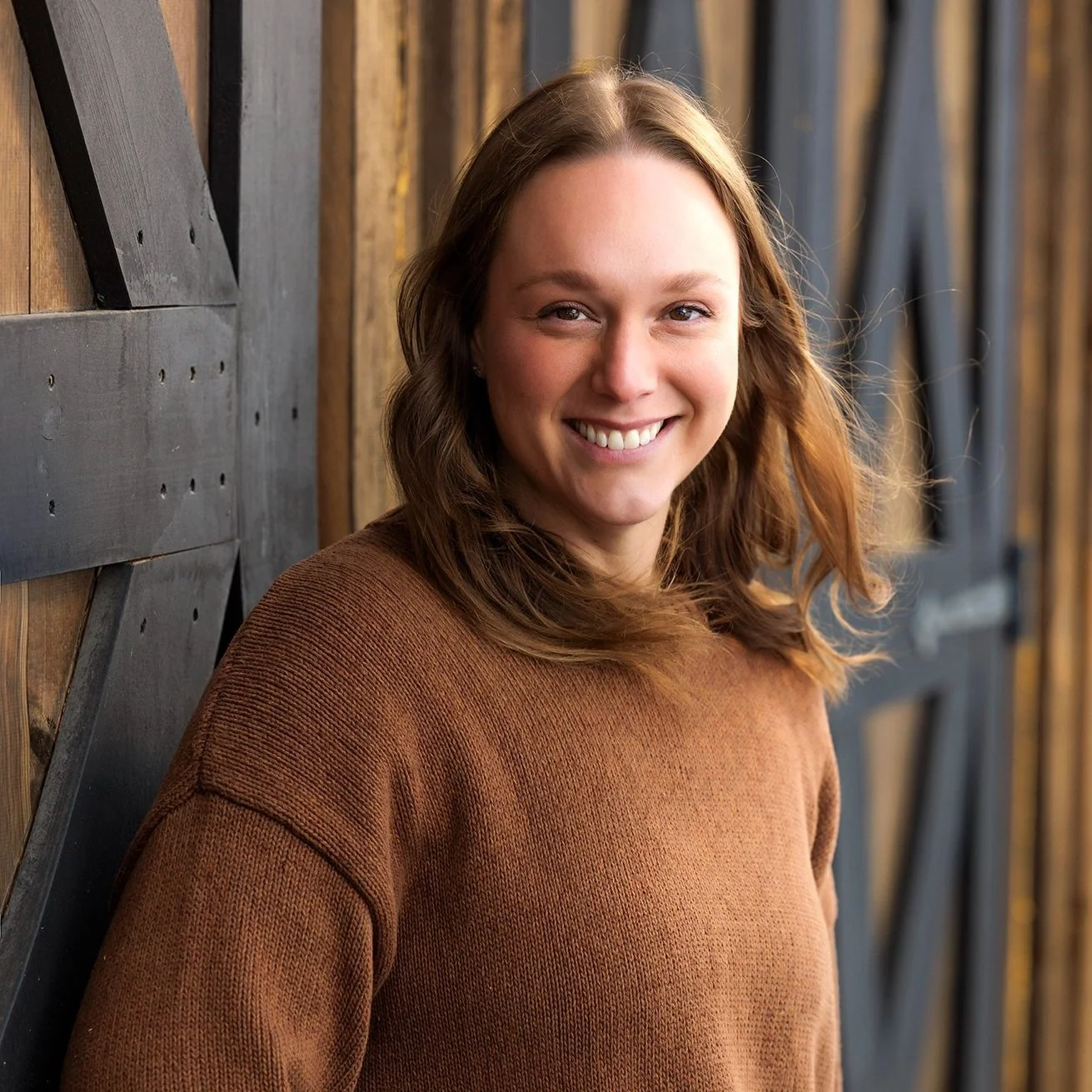 A smiling young woman with shoulder-length brown hair, wearing a brown sweater, standing against a wooden and black geometric wall.