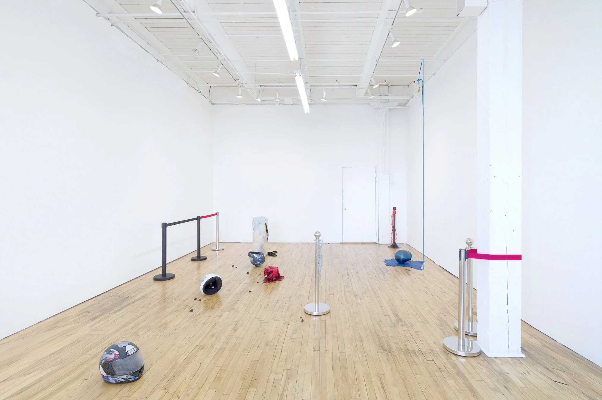 Empty art gallery room with scattered miscellaneous objects, including a helmet on the floor, barrier ropes, and a helmet, with white walls, wooden floor, and ceiling lights.