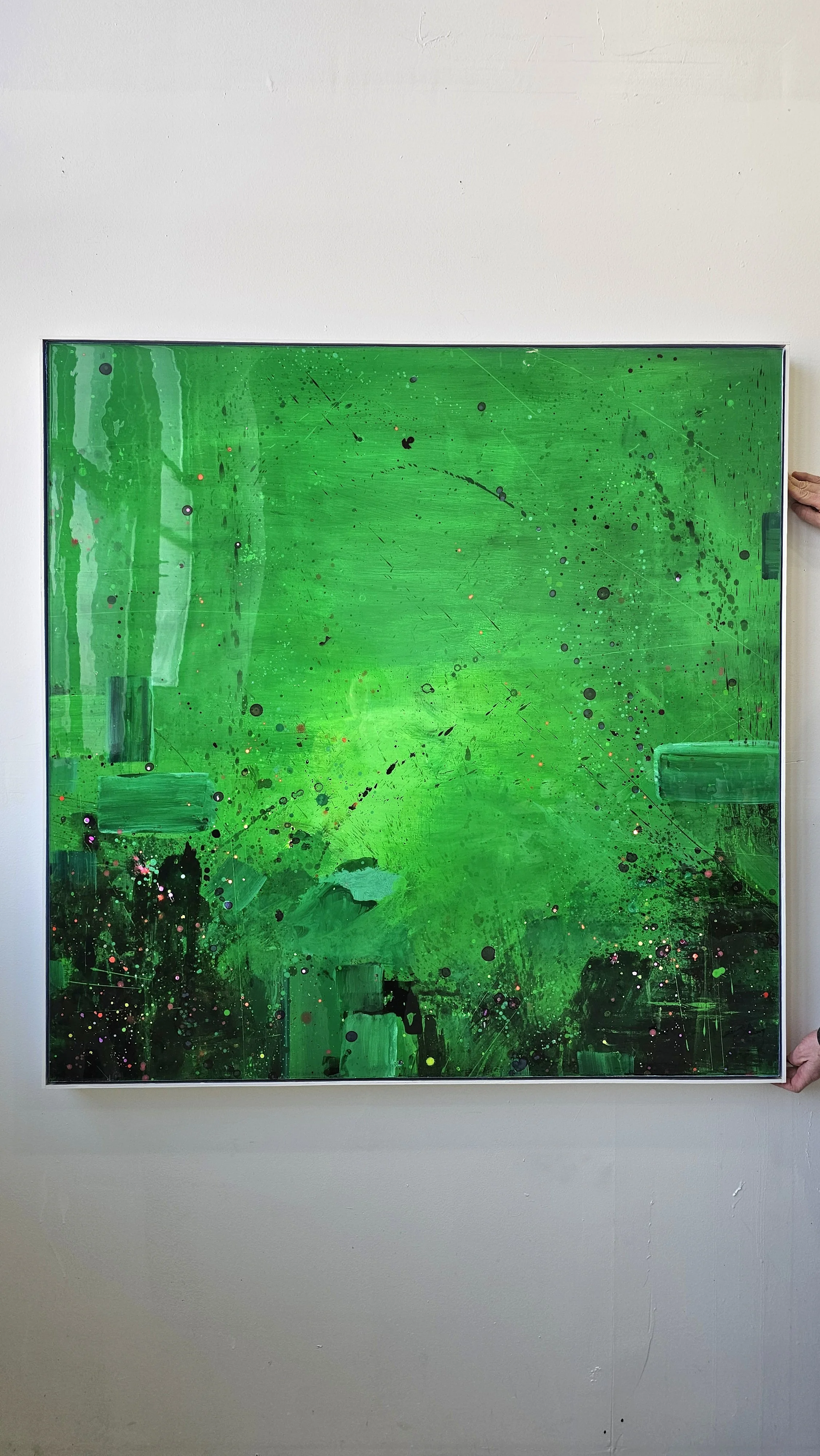 Abstract green painting with splashes and streaks, framed and displayed against a white wall, with a person's hand visible on the right side holding the frame.