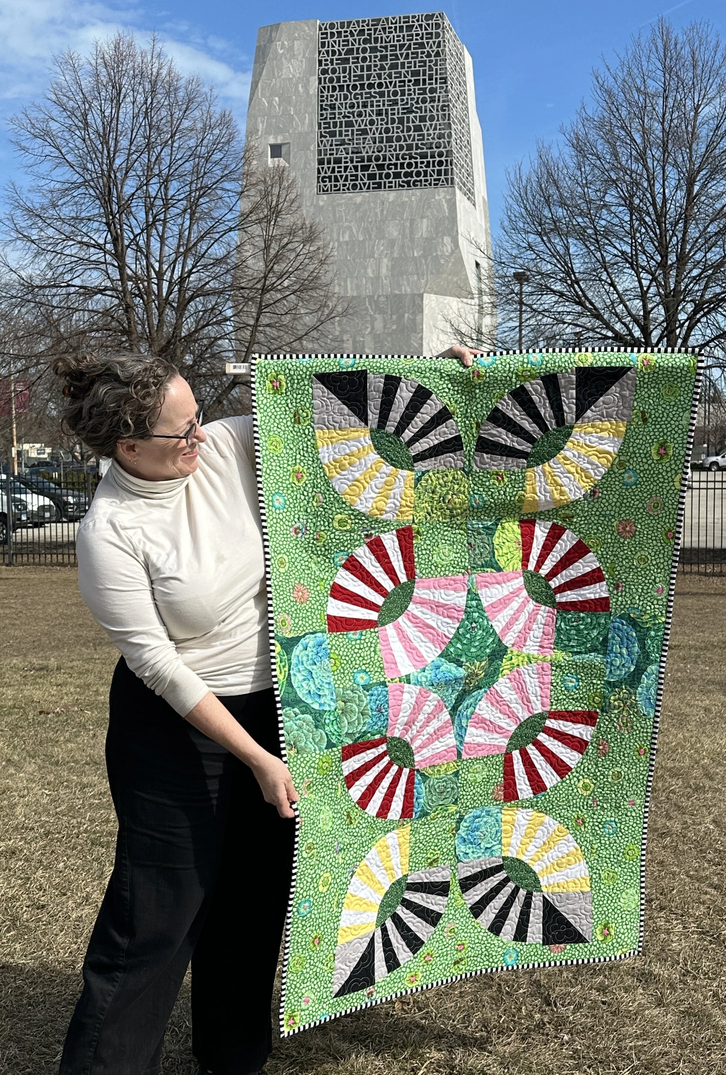 A woman standing outside holding a colorful quilt with geometric patterns. The quilt has large circular designs in various colors, including pink, green, black, white, yellow, and red, set against a green background with small floral patterns. In the background, there are leafless trees, a parking lot, and a tall modern building with a large text inscription on its upper side against a partly cloudy sky.