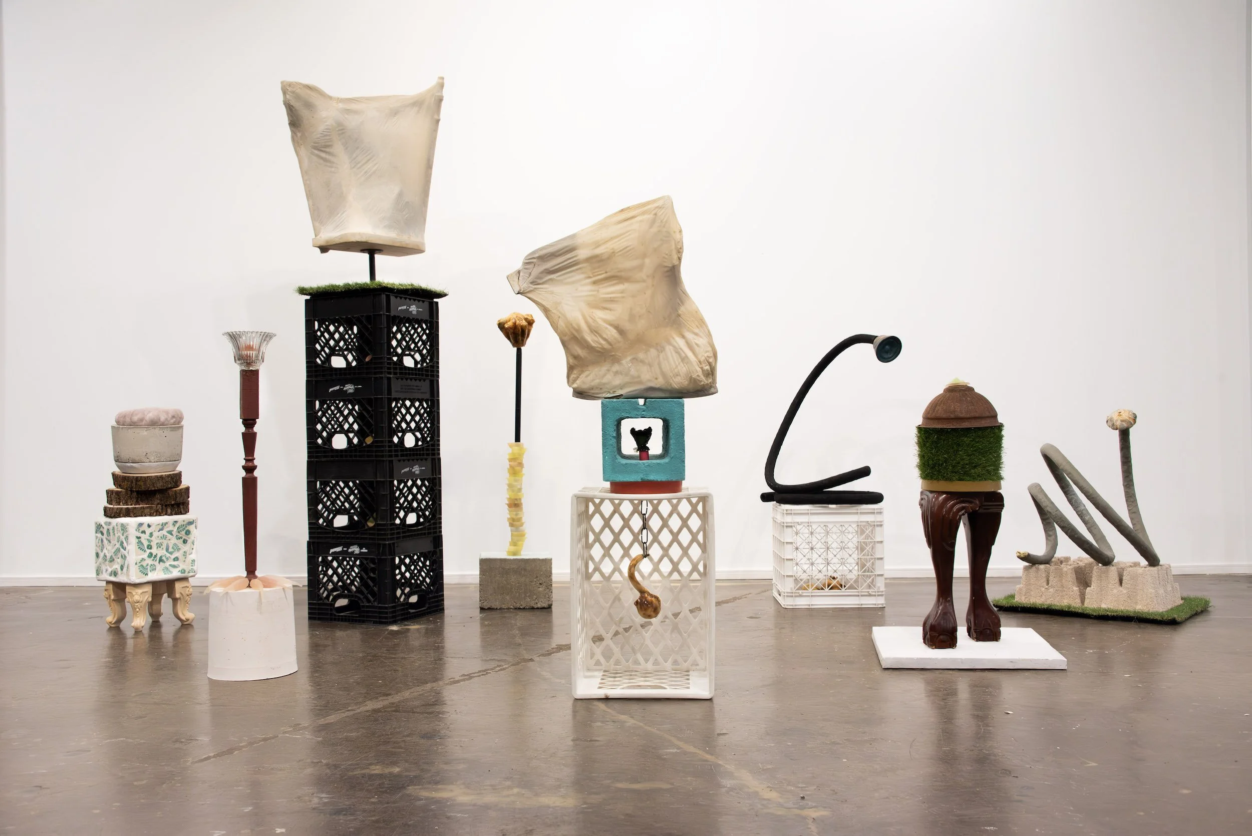 Art installation or sculpture display with various abstract and unconventional objects arranged on different platforms and stands against a plain white wall.