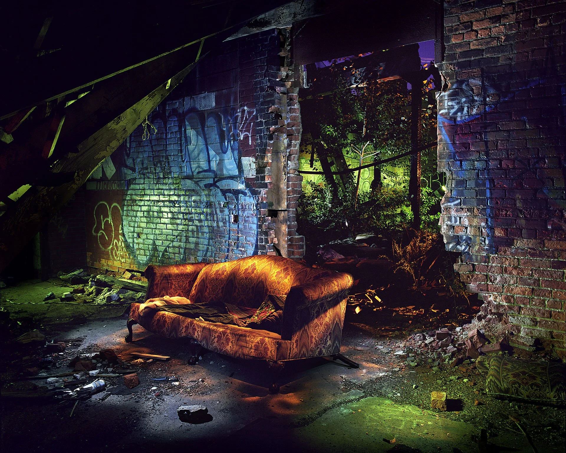 Dilapidated room with exposed brick walls and a torn sofa. Graffiti on the walls, rubble on the floor, and debris scattered around. Green and purple lighting create a moody atmosphere.