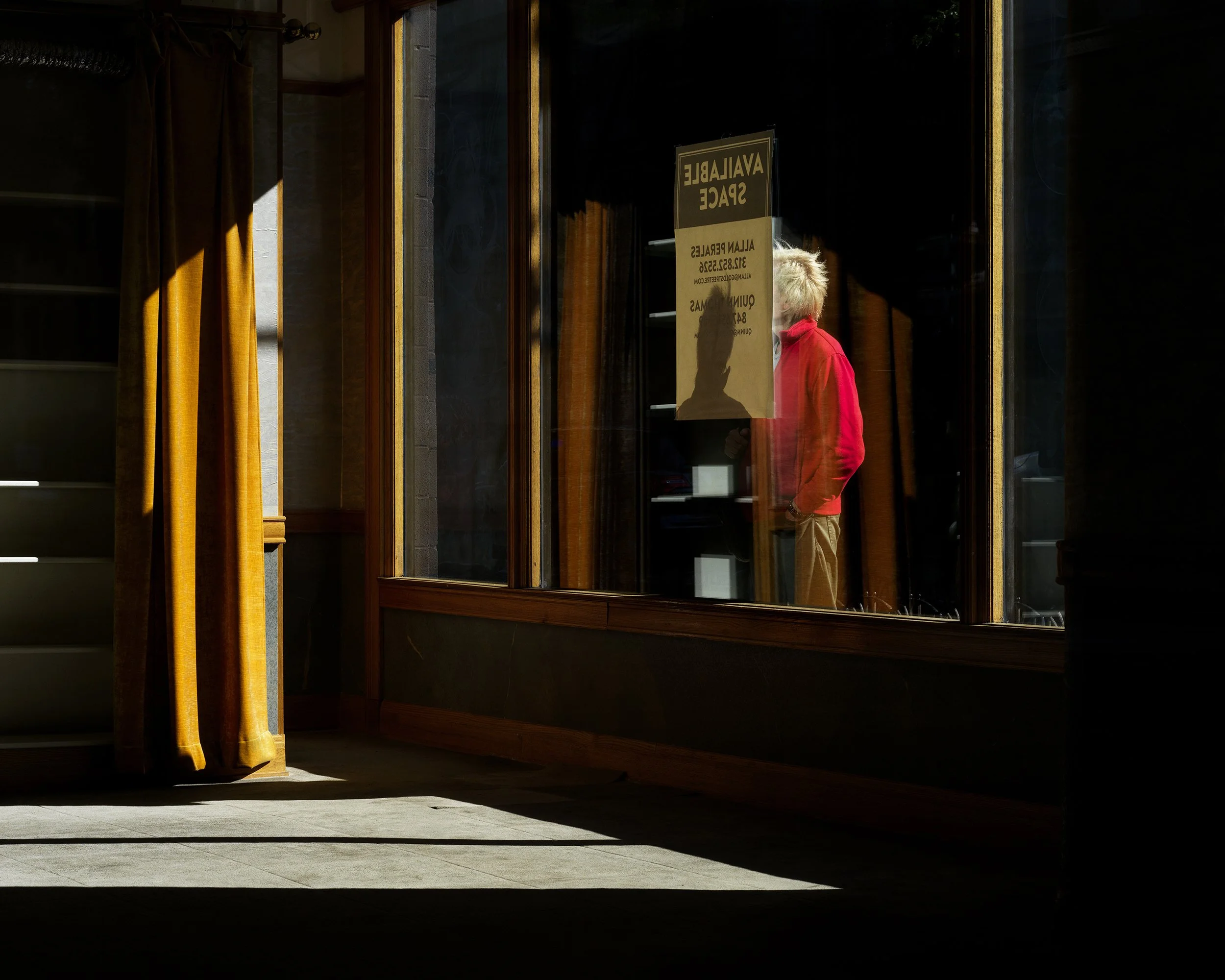 A person with blonde hair wearing a red jacket inside a building, visible through a glass door that has a sign on it.
