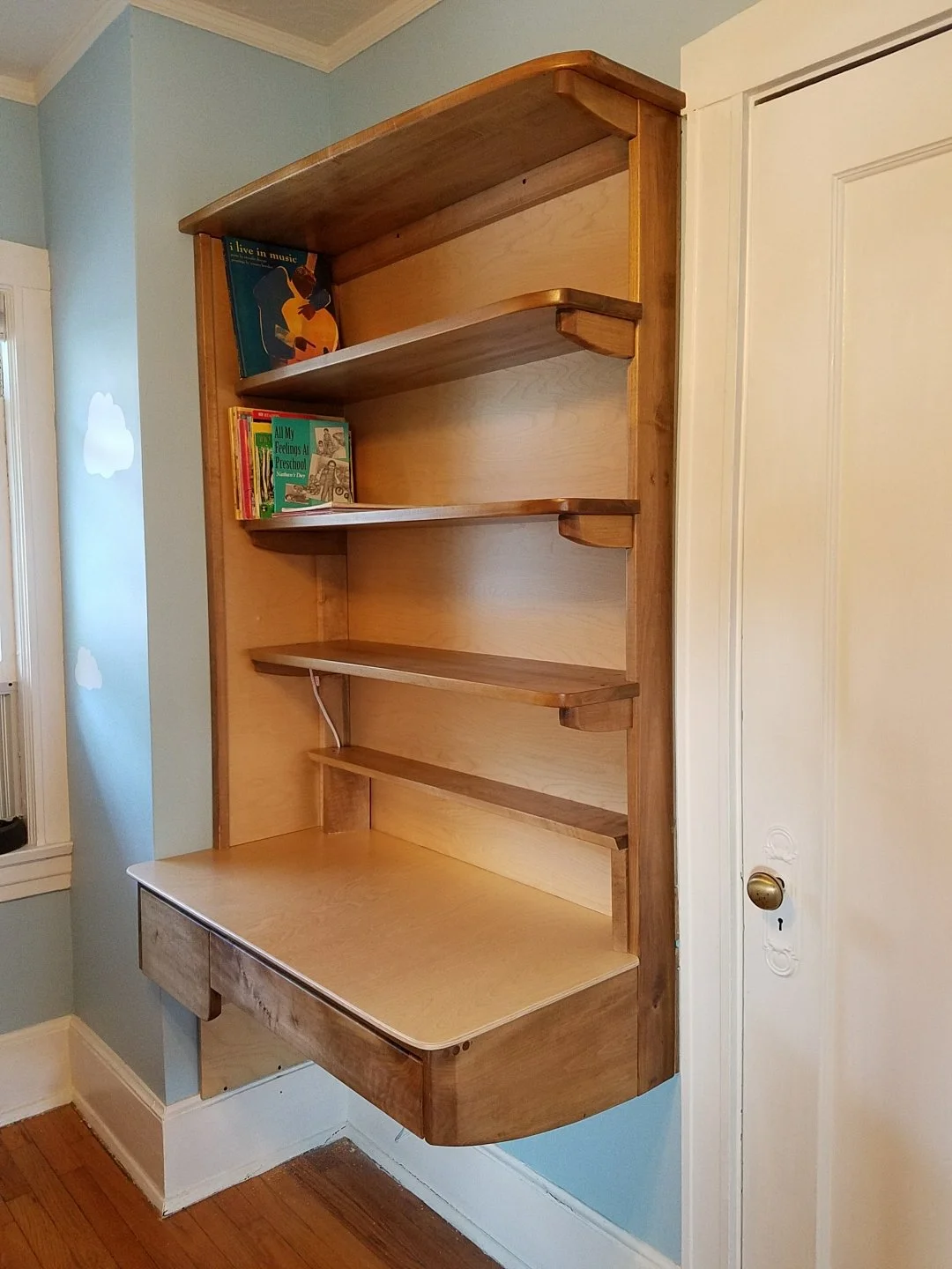 Wooden wall bookshelf with multiple shelves and a lower pull-out desk, situated near a white door and light blue wall with cloud decals.