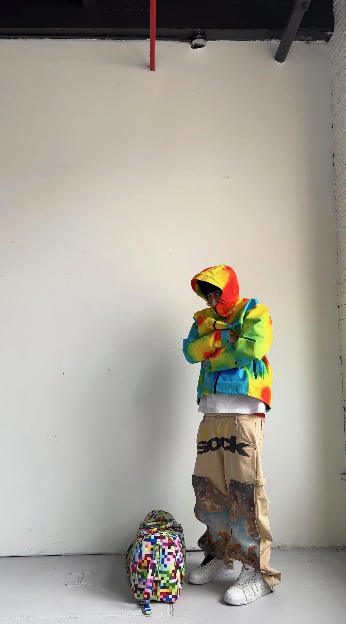 Person standing against a white wall wearing a colorful tie-dye hoodie with the hood up, beige baggy pants with patches, and white sneakers. There is a pixelated multicolored backpack on the floor in front of them.
