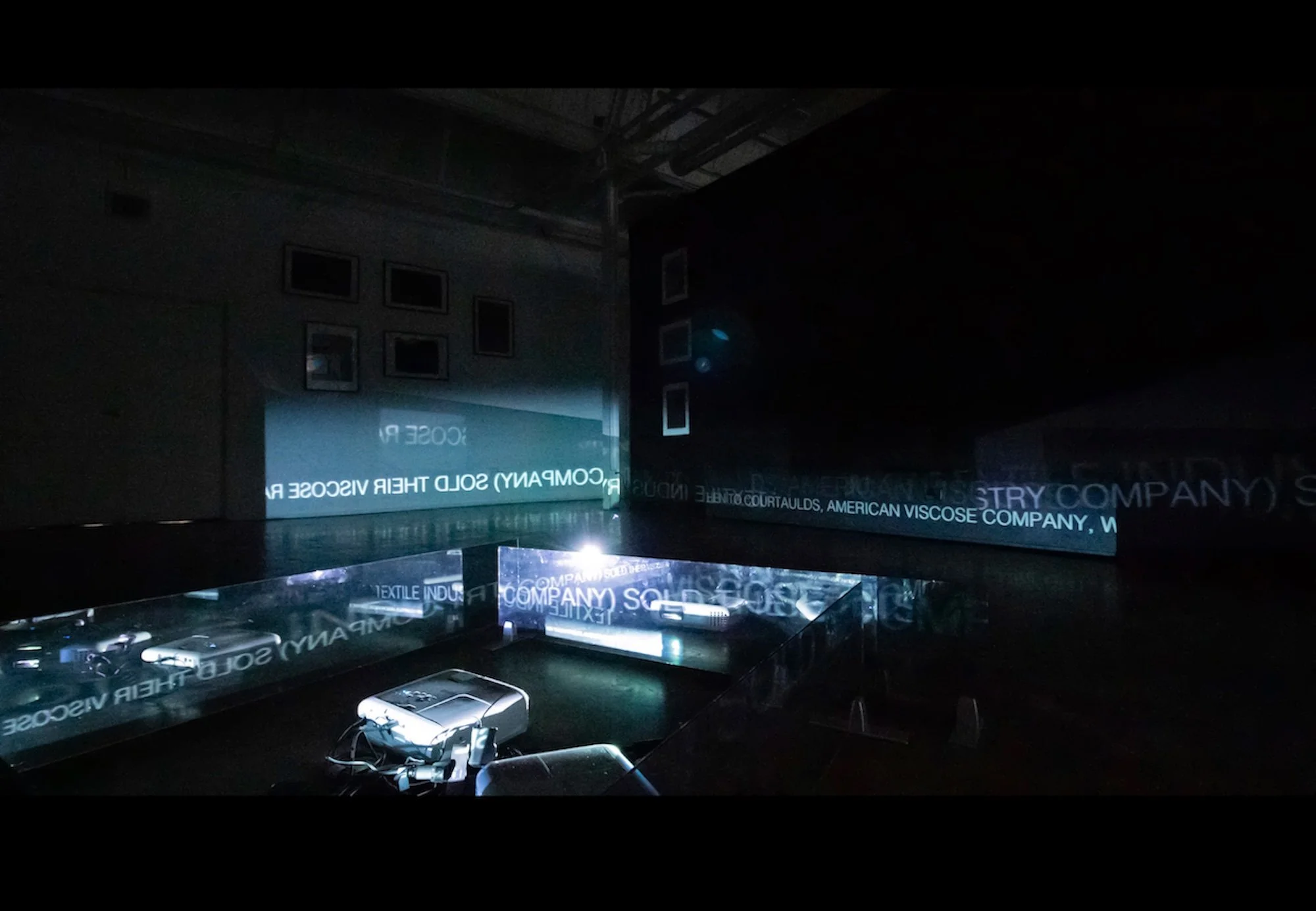 Dimly lit room with posters and screens, illuminated by projector light, displaying texts about American viscose and textile industries.