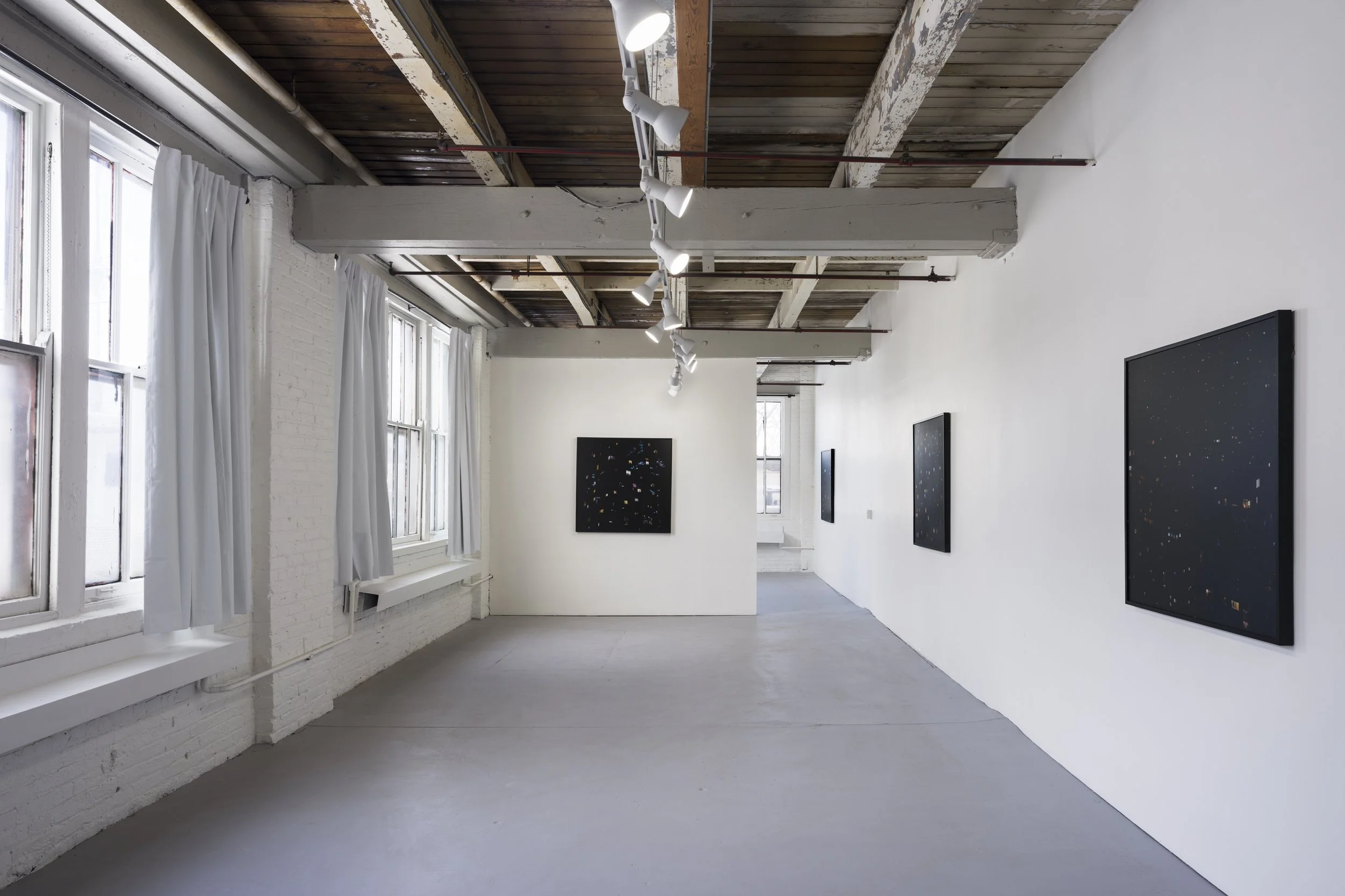 An art gallery with white painted brick walls, large windows with white curtains, and a ceiling with exposed wooden beams and track lighting. There are black framed art pieces hanging on the walls.