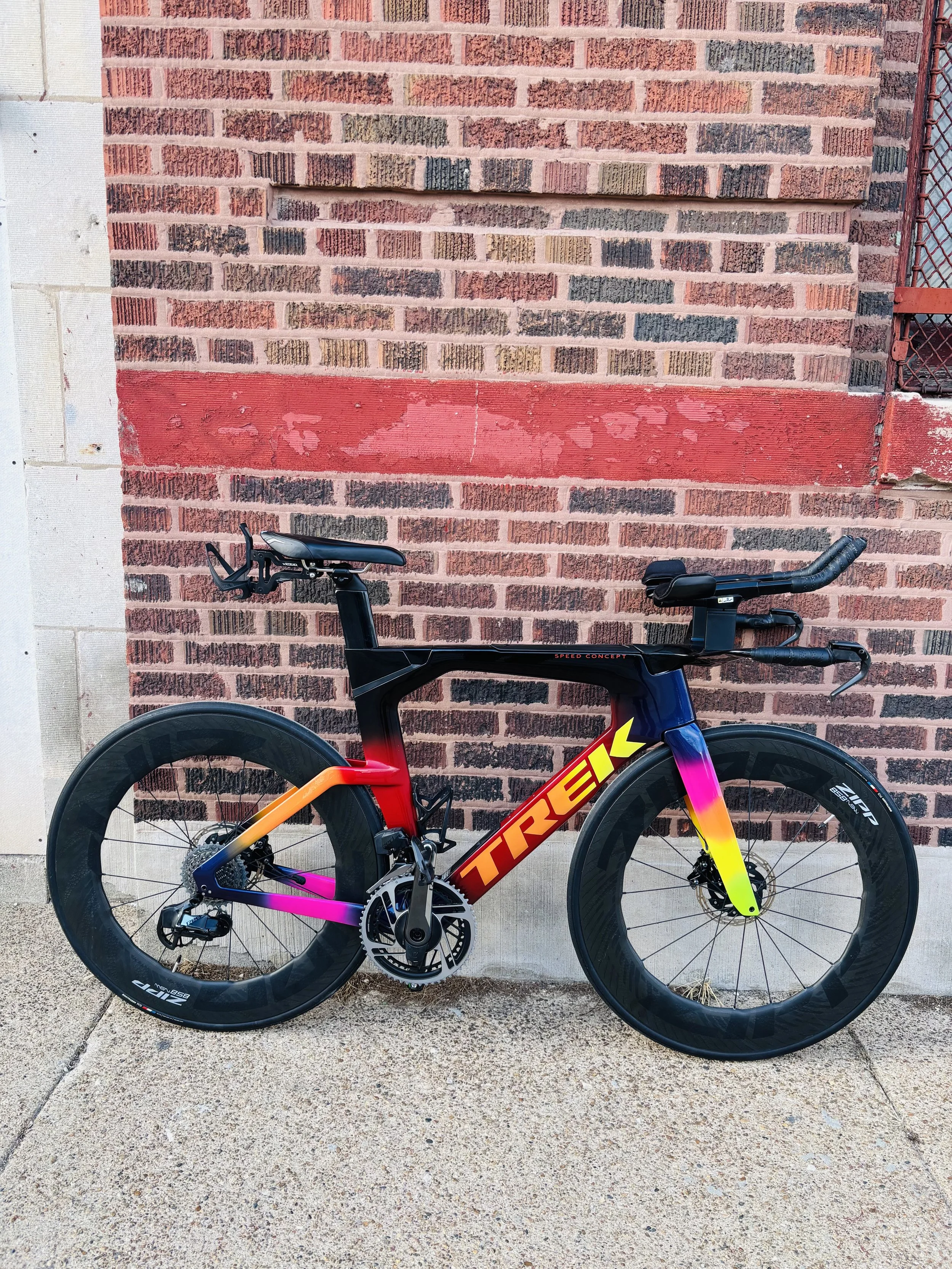 A black and yellow Trek racing bicycle with red and pink accents leaning against a brick wall with a red stripe. The bicycle has deep-section wheels, disc brakes, and flat handlebars.