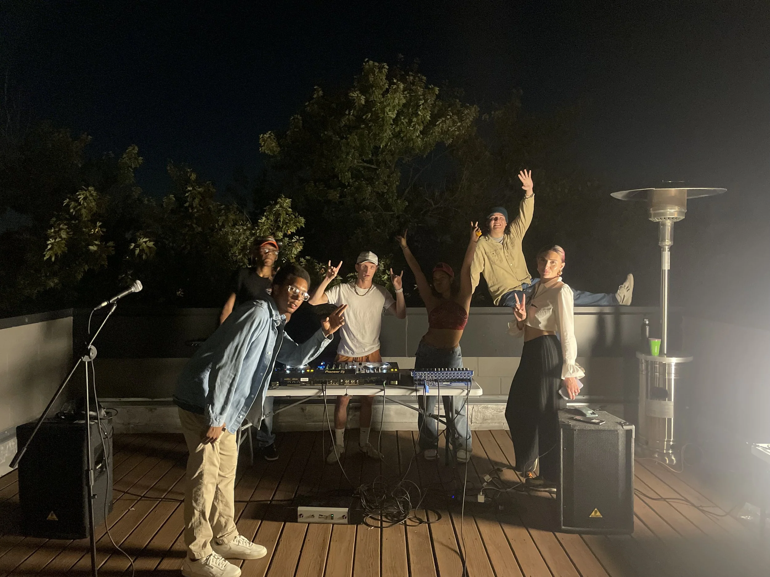 A group of people at an outdoor party or event, some making peace signs, with DJ equipment, a microphone, and a patio heater, at night under trees.