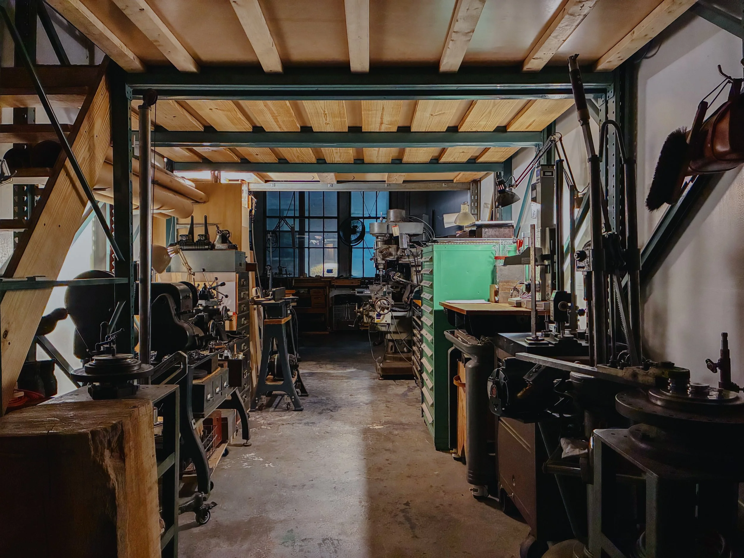 A workshop with woodworking tools, machines, and workbenches, with a high ceiling featuring wooden beams and a large window at the back.