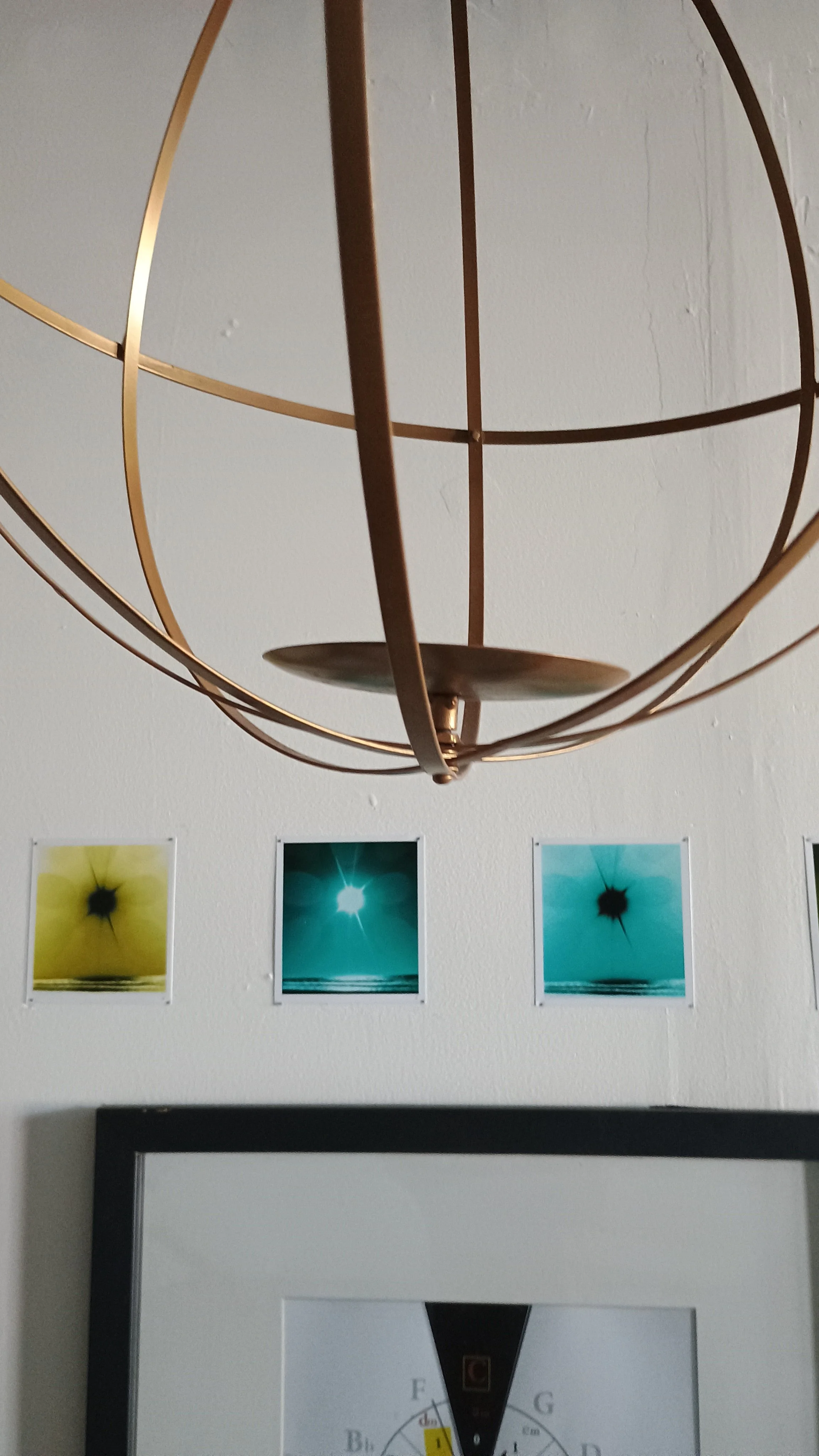 A modern chandelier with a spherical metal frame hangs from the ceiling. Below it, on the wall, are four colorful abstract photographs arranged in a row.